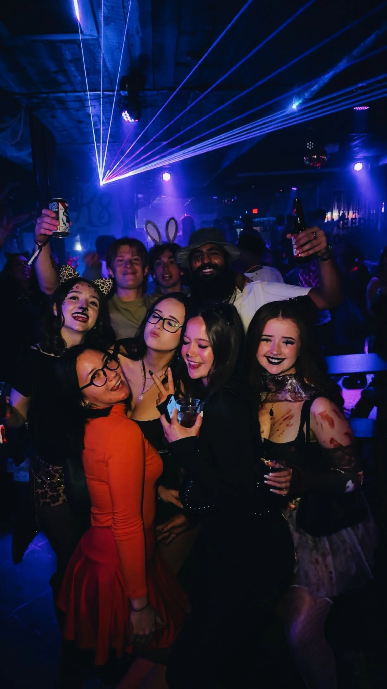 Group of people dressed in costumes at a Halloween party, with colorful lighting and laser effects in the background.