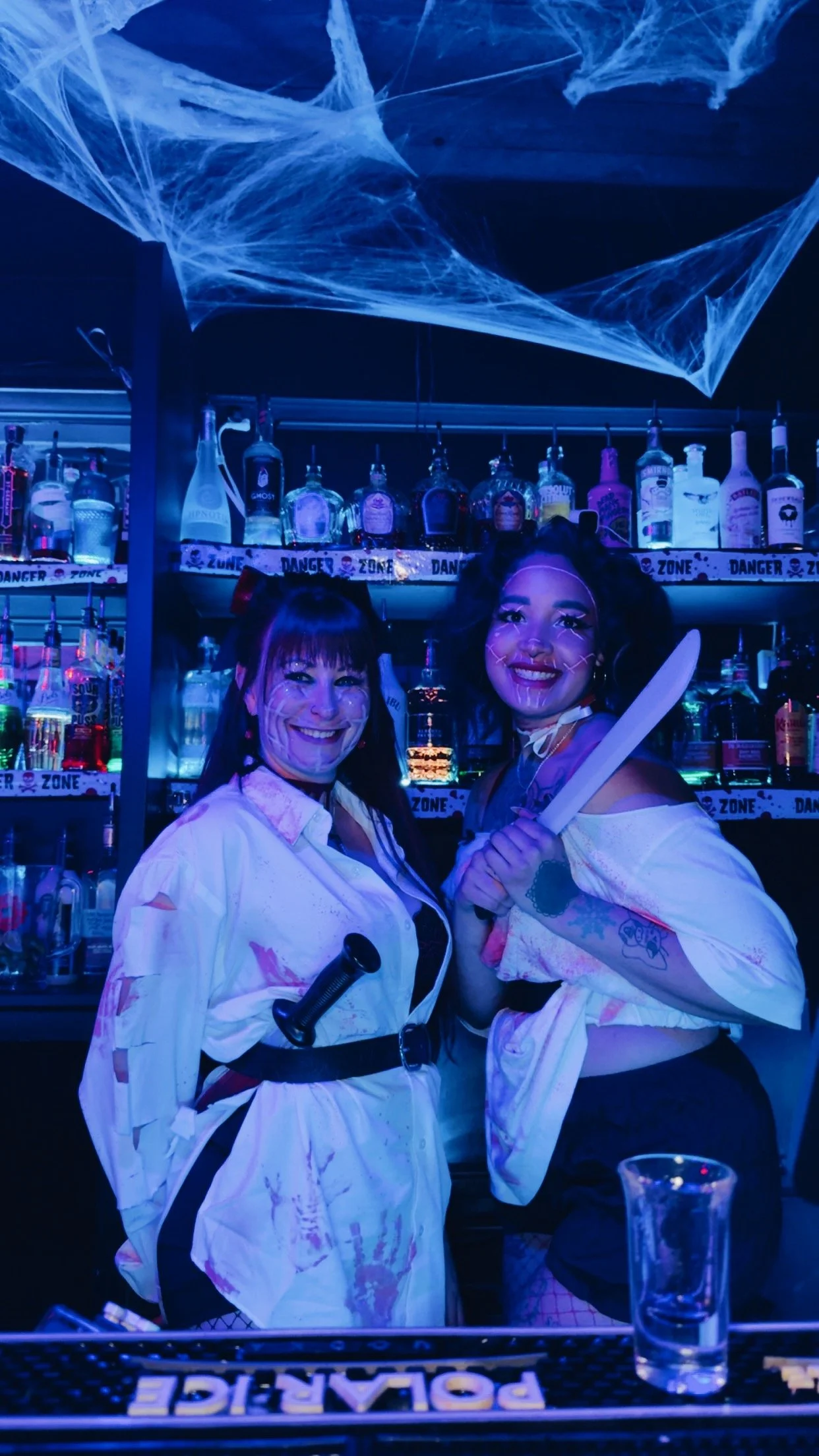 Two women dressed as horror movie characters standing behind a bar, smiling and holding a machete, decorated with Halloween props and blue lighting.