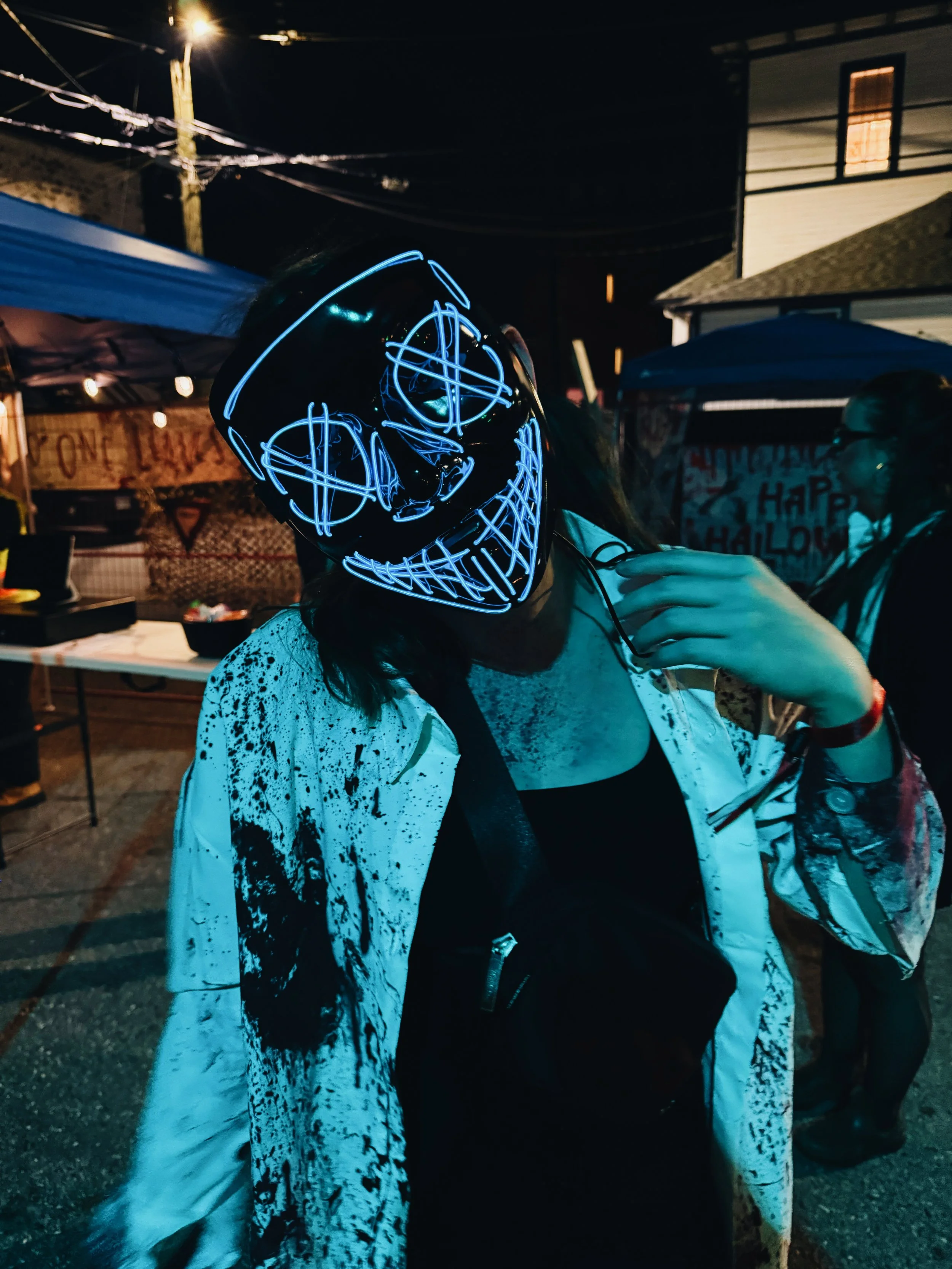 Person wearing a glowing LED Halloween mask with a creepy face design, standing outdoors at night during a Halloween celebration, with tents and other people in the background.