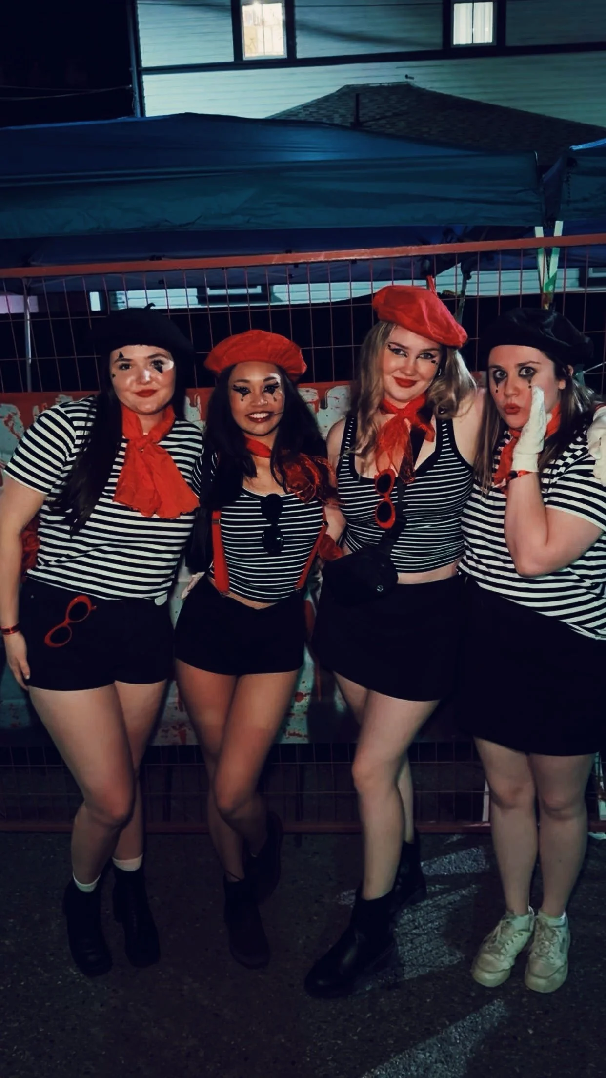 Four women dressed in French mime costumes standing side by side, wearing black and white striped shirts, black skirts, red scarves, berets, and some with theatrical makeup.