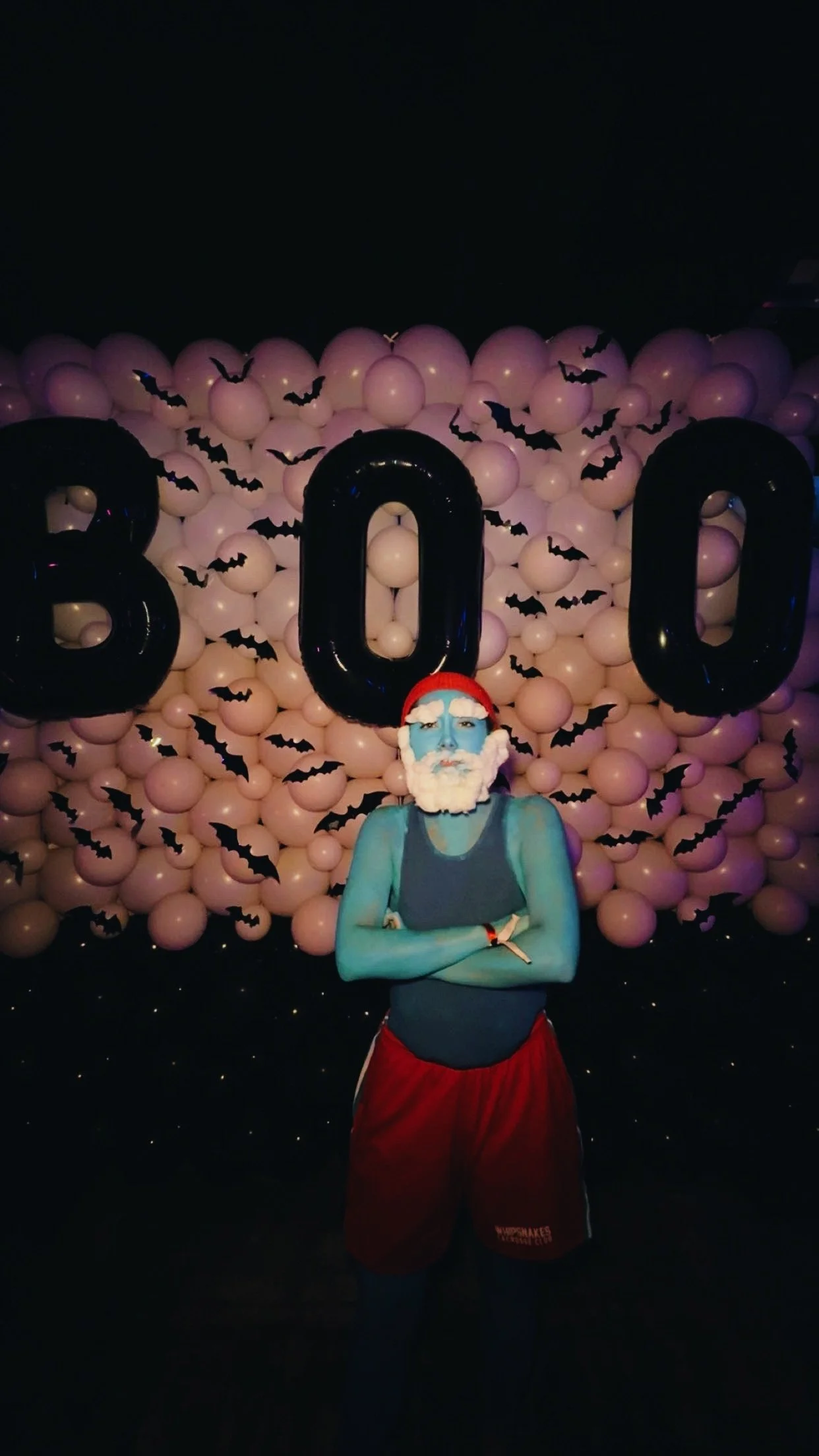 Person with blue face paint and Santa hat standing with arms crossed in front of a Halloween-themed balloon backdrop with the numbers 2020 and bat decorations.