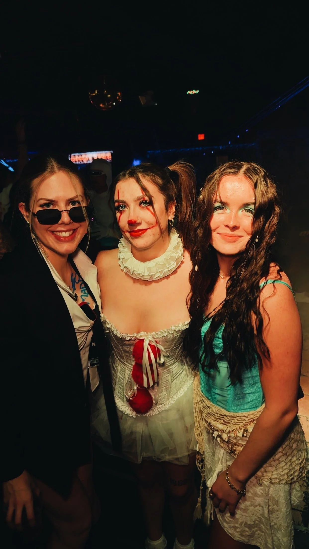 Three women dressed in costumes at a party, one with sunglasses and a suit, another as a clown, and the third in a colorful dress with long wavy hair.