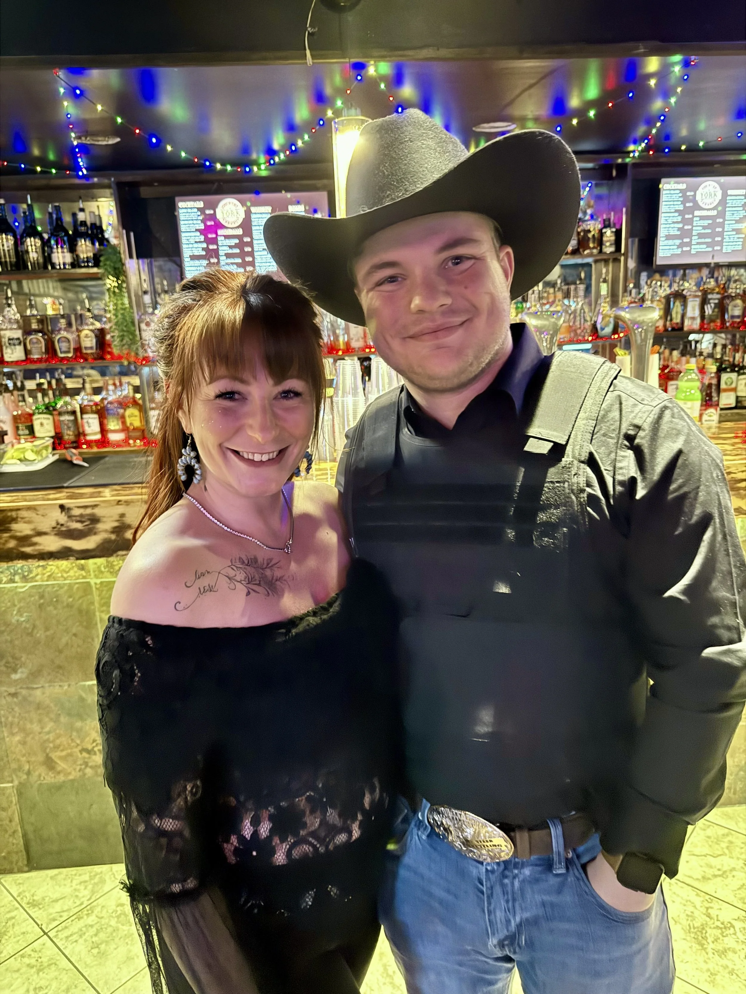 A smiling woman with tattooed shoulders and earrings standing next to a smiling man in a cowboy hat at a bar with colorful string lights and illuminated screens in the background.