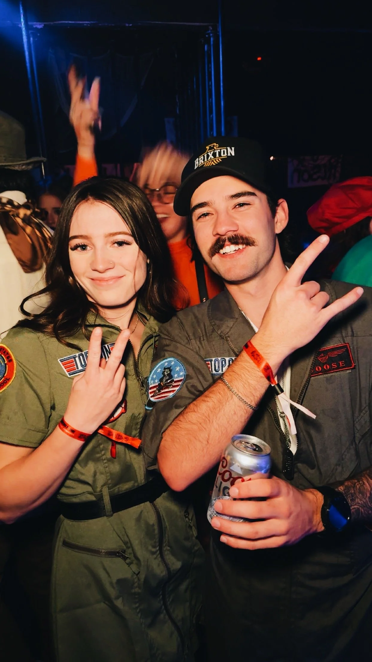 Two young people in military-style costumes at a party, making peace and rock hand signs, with one holding a can of soda.
