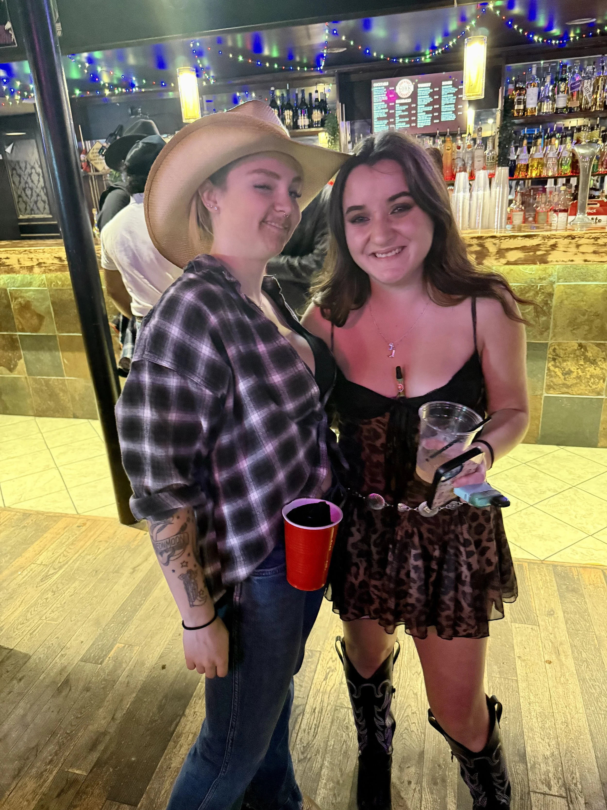 Two women smiling at a bar, one wearing a large cowboy hat and plaid shirt, and the other in a black, leopard print dress with cowboy boots. They are holding drinks and posing together.
