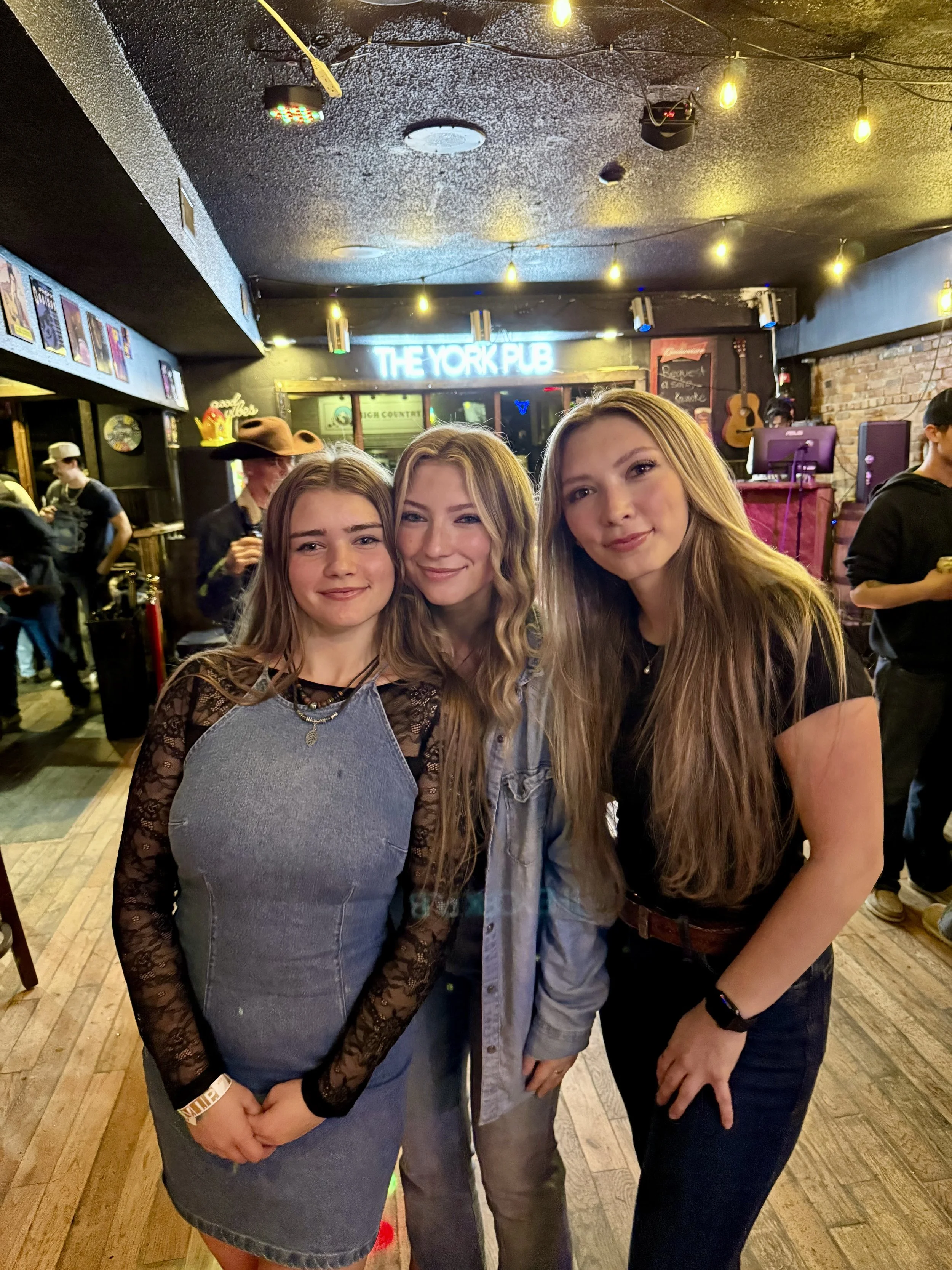 Three young women smiling and posing together in a bar or pub with a neon sign that says 'The York Pub' in the background. The setting has warm lighting and a casual, lively atmosphere.