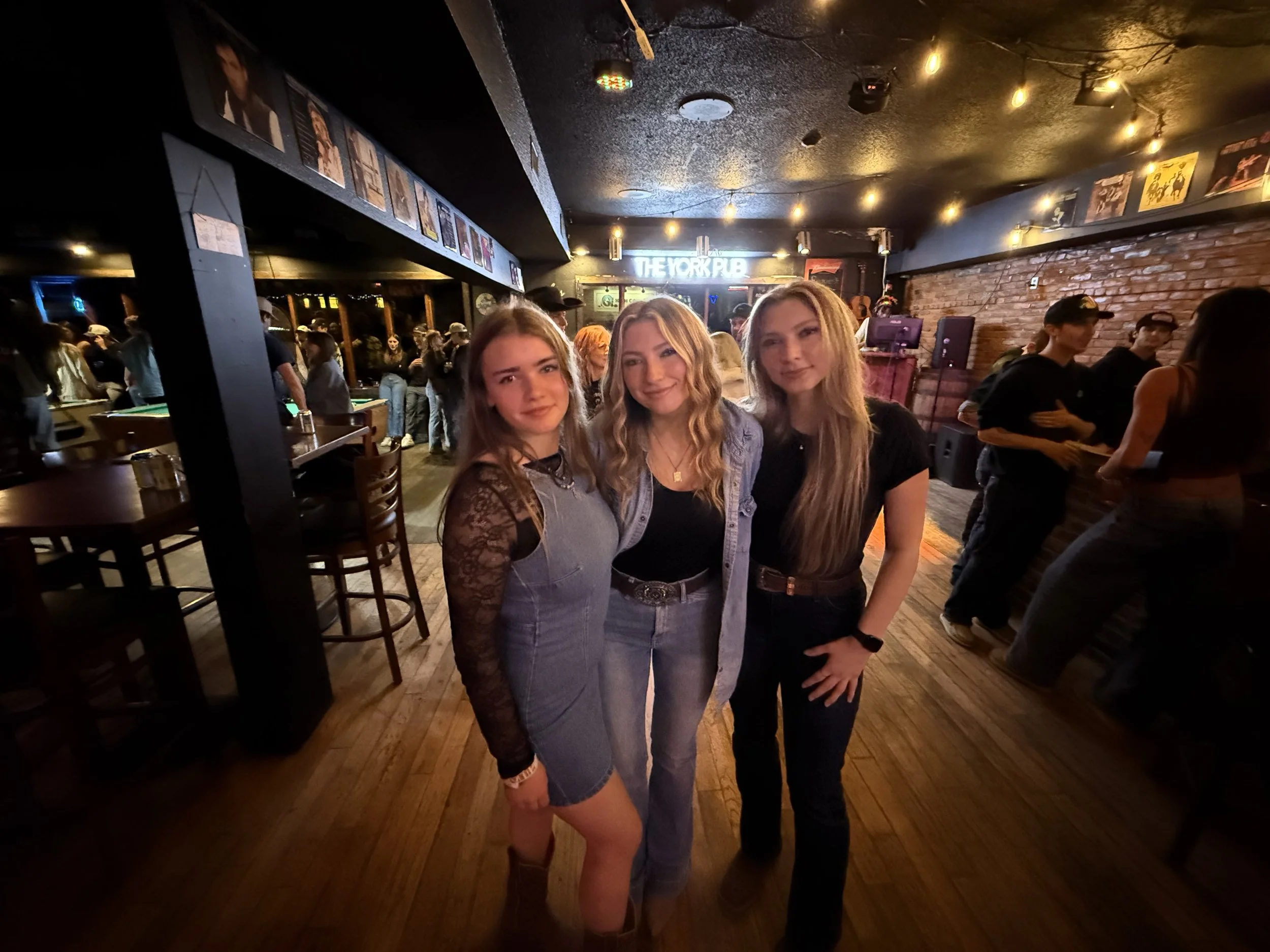 Three young women standing together in a dimly lit bar or music venue, with other people in the background. The woman on the left is wearing a gray dress with lace sleeves, the woman in the middle is wearing a light denim jacket over a black top, and