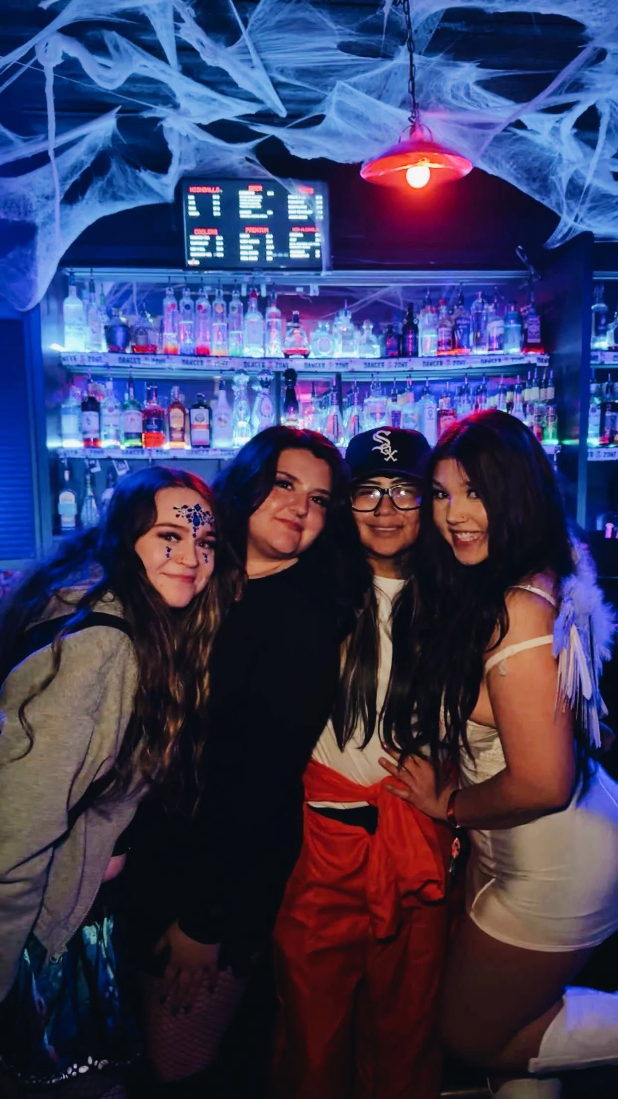 Four women standing in front of a bar with illuminated bottles, decorated with fake cobwebs and illuminated by red and purple lights, at a Halloween-themed party.