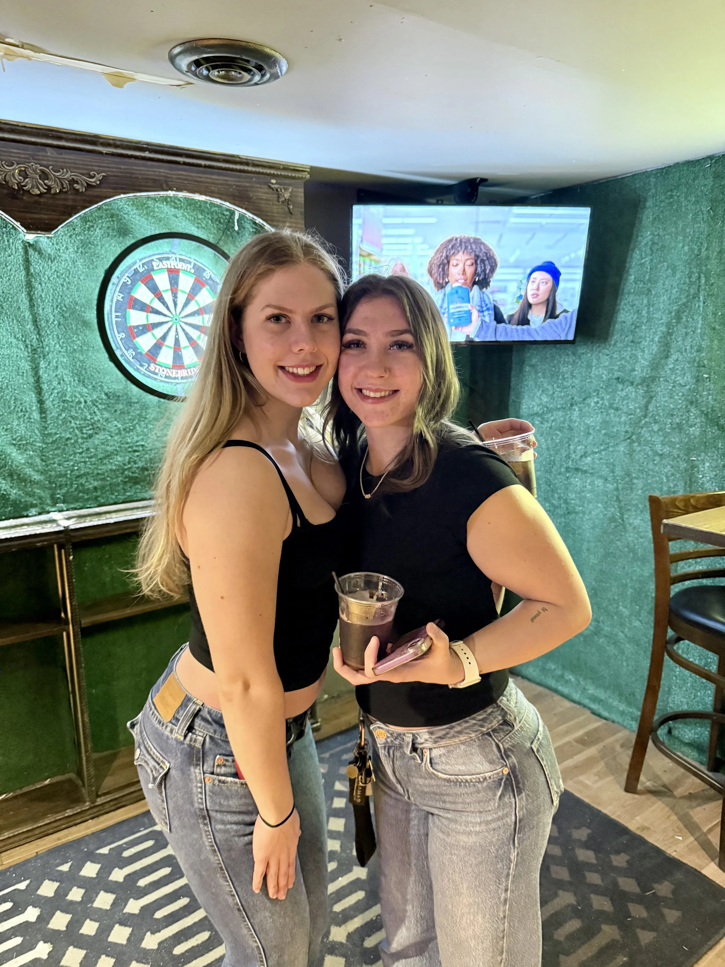 Two young women standing close together in a bar, smiling at the camera, holding drinks in plastic cups, with a dartboard and a television screen in the background.