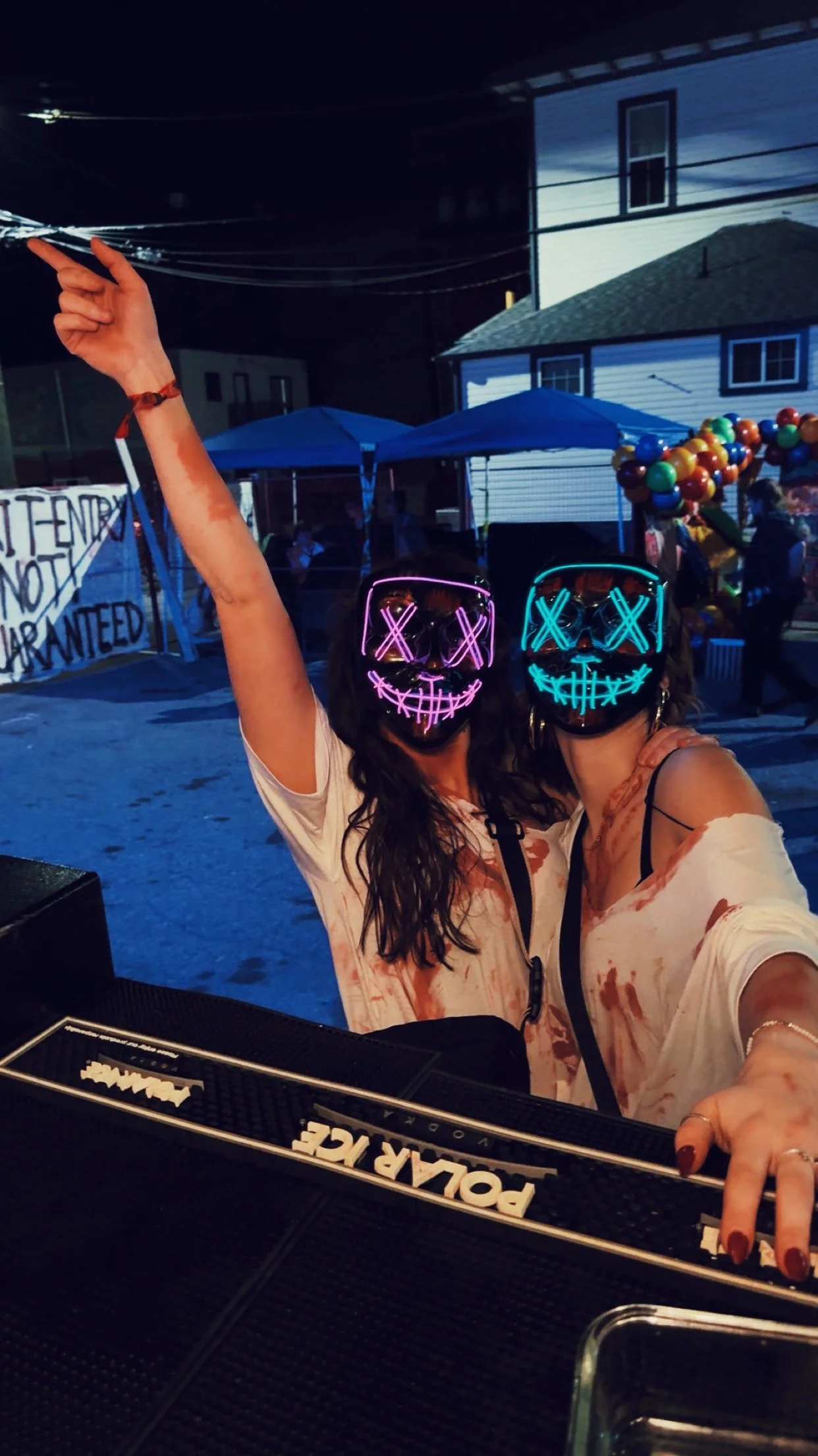 Two women wearing illuminated skull masks, posing for a photo at a nighttime party or event with tents, balloon decorations, and a house in the background.
