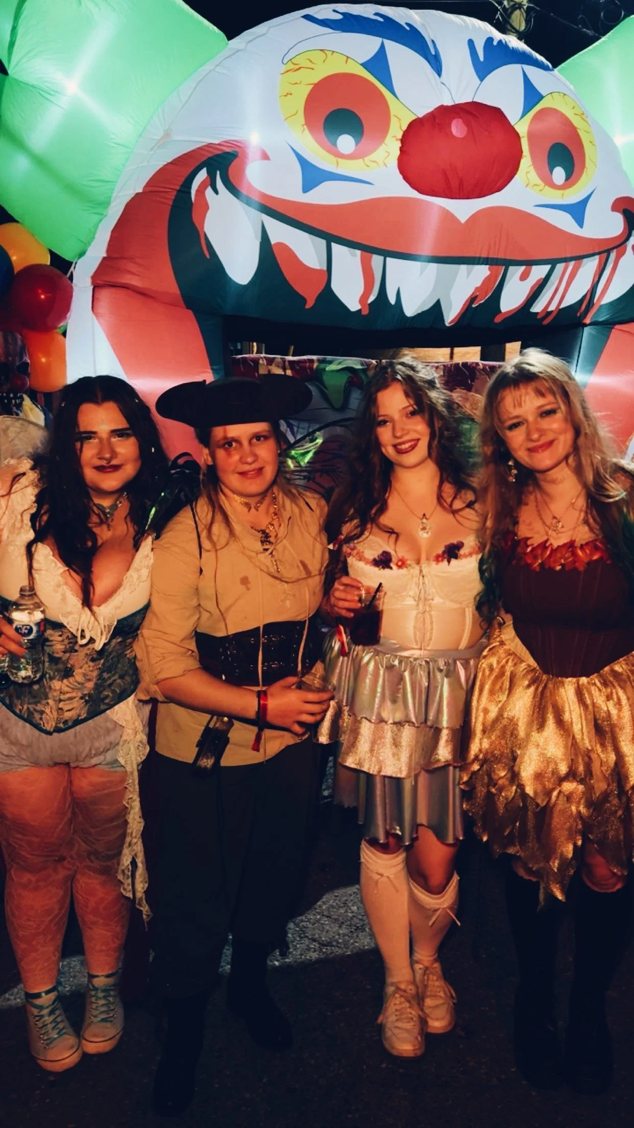 Four women standing together at a costume party, with a large colorful clown face balloon in the background.