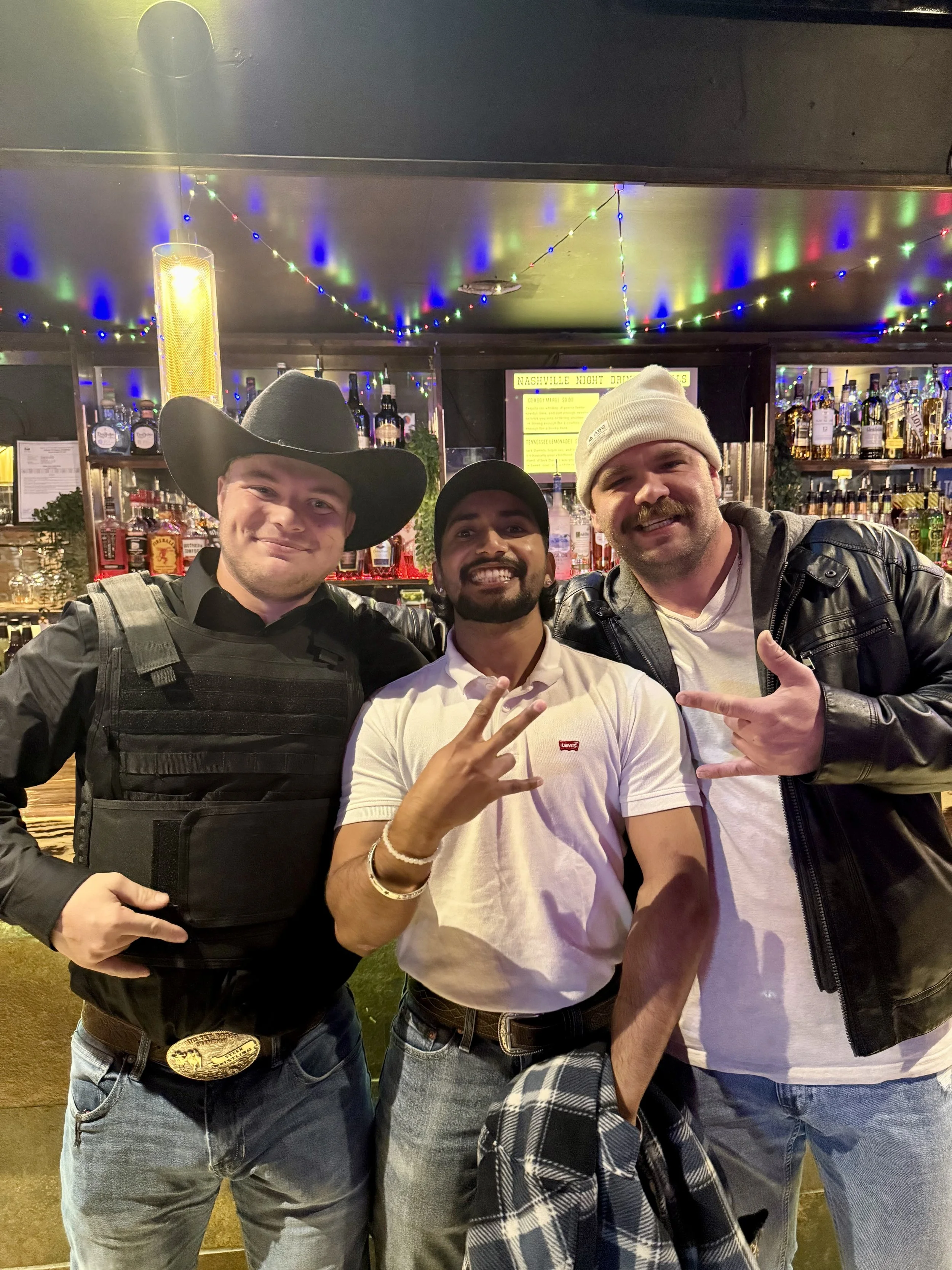 Three smiling men posing together in a bar, with bottles of alcohol and colorful string lights in the background. The man on the left wears a cowboy hat and a tactical vest, the man in the middle wears a baseball cap and a white shirt, and the man on