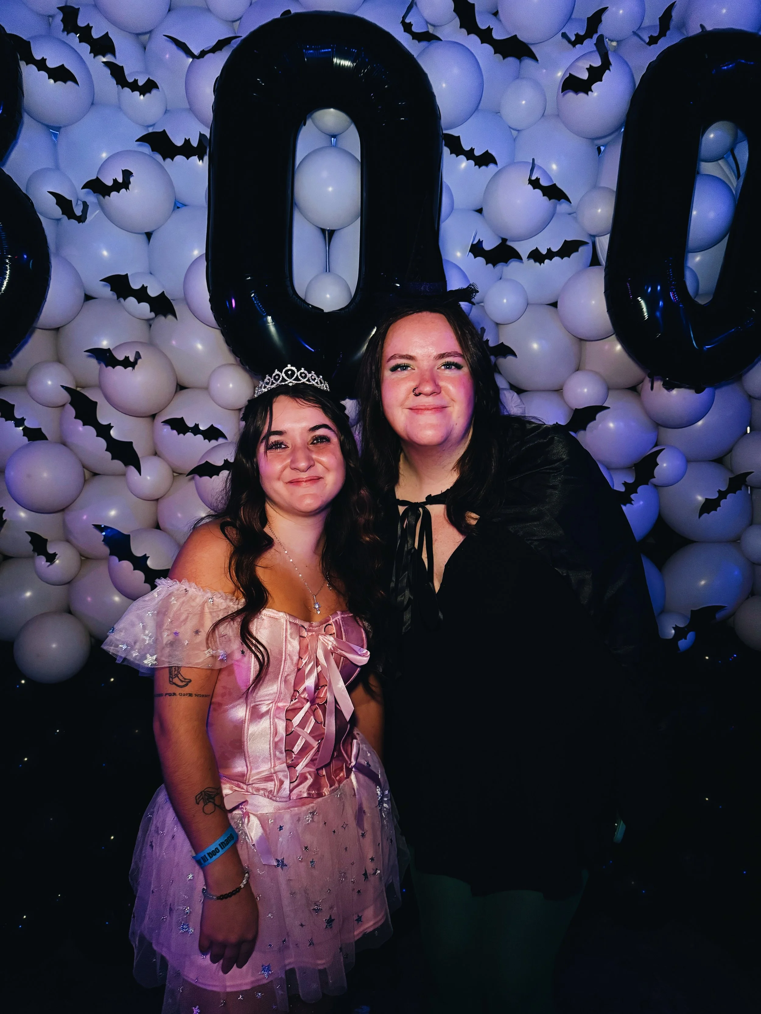 Two women dressed in costume pose in front of a Halloween-themed background with balloon letters and bats.