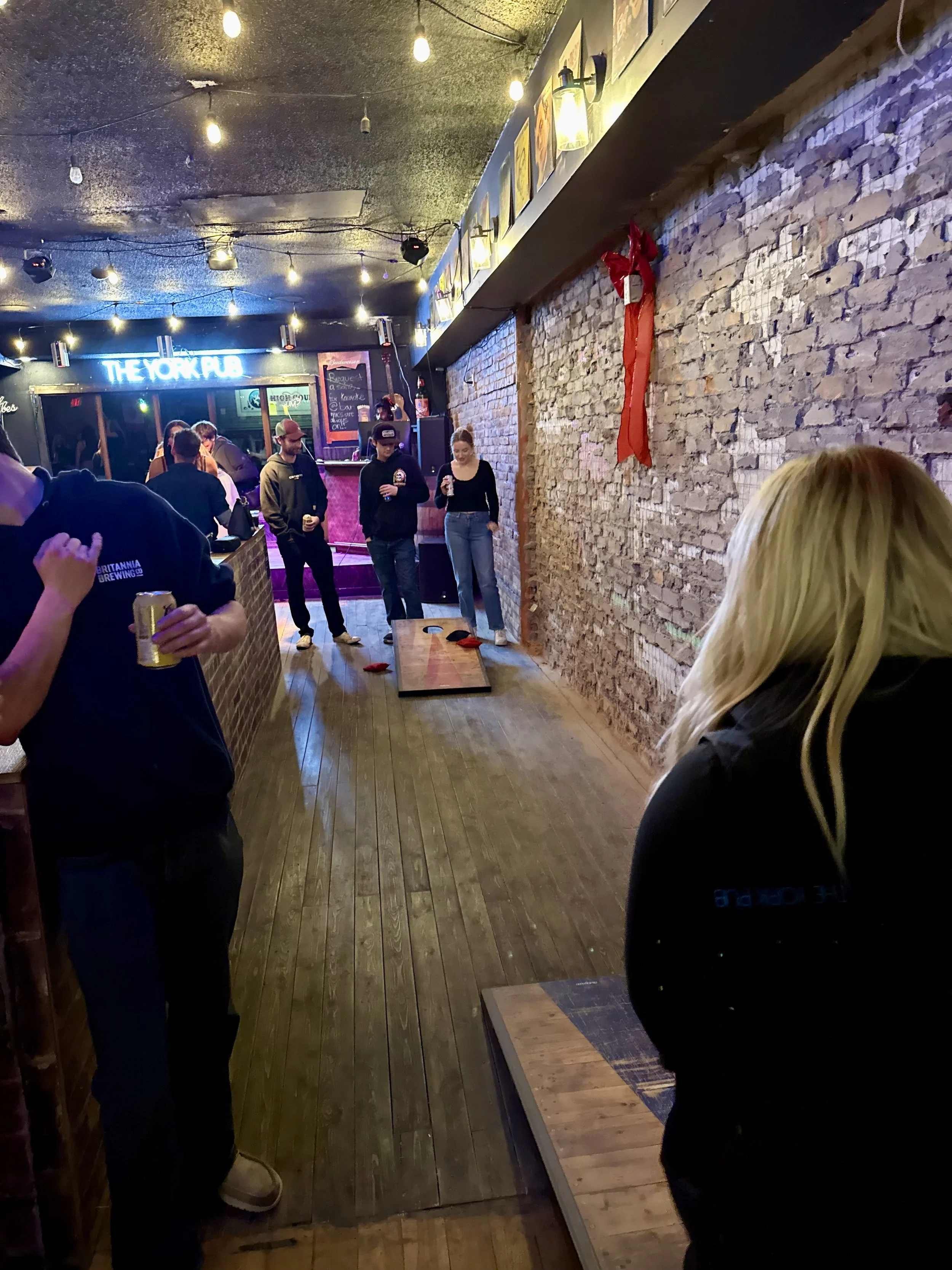 People playing cornhole in a bar with exposed brick walls, string lights, and a neon sign that says 'The York Pub.'