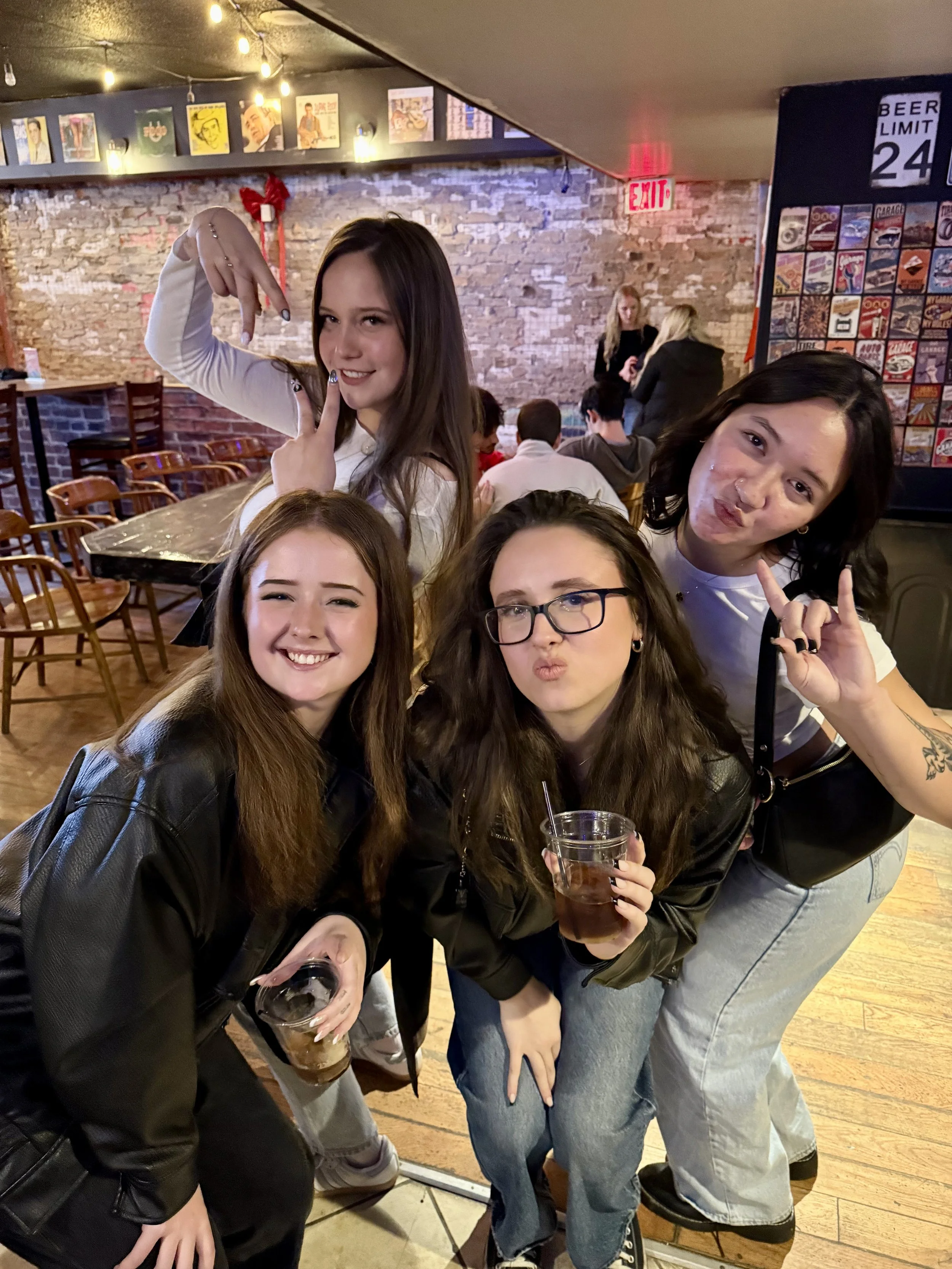 Four women posing and smiling at a bar or restaurant with exposed brick wall and framed artwork, with people sitting in the background.