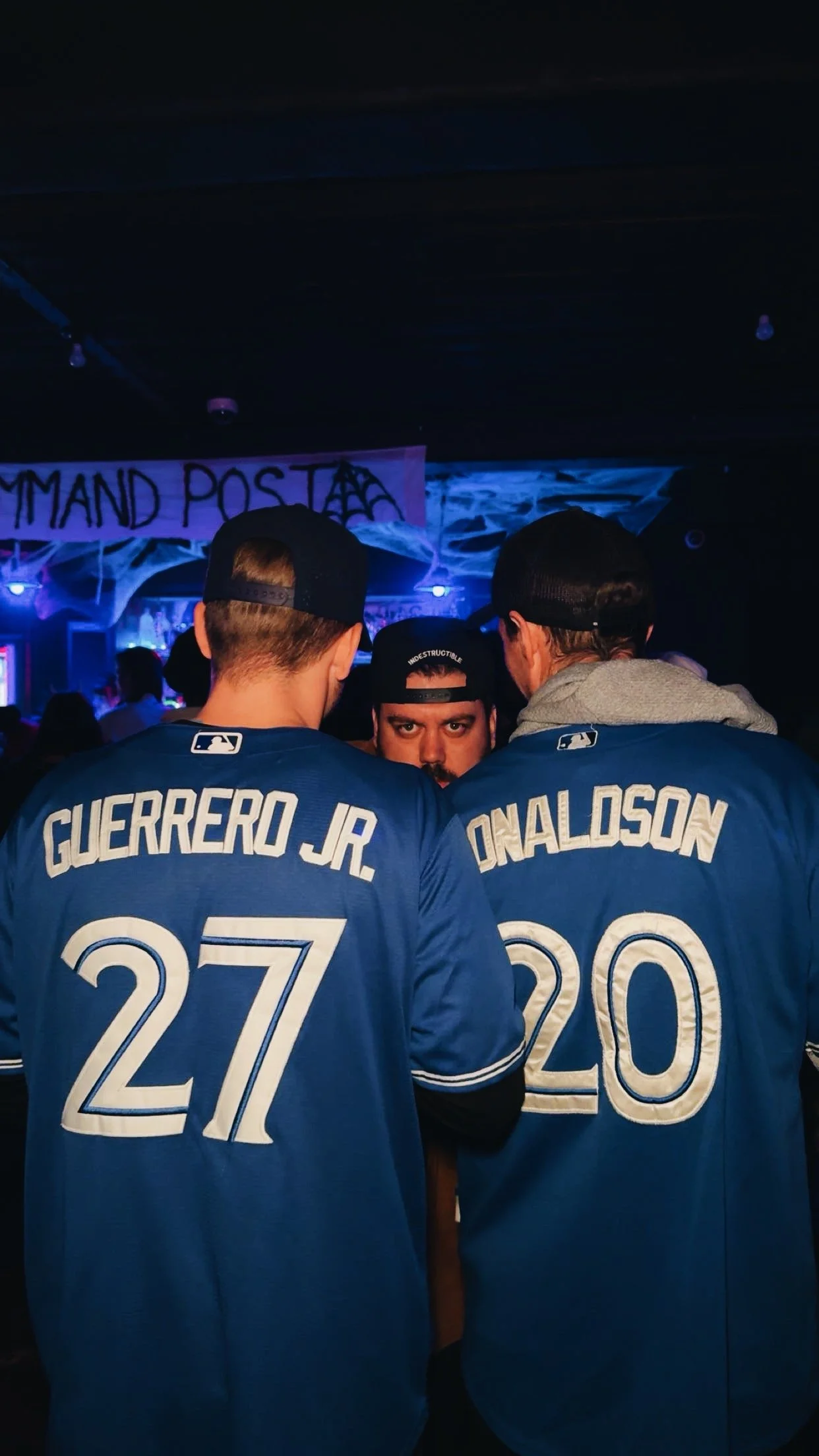 Two men in blue baseball jerseys with the names Guerrero Jr. and Onalosa and the numbers 27 and 20 on their backs, standing with a person in the background in a dark, decorated room.