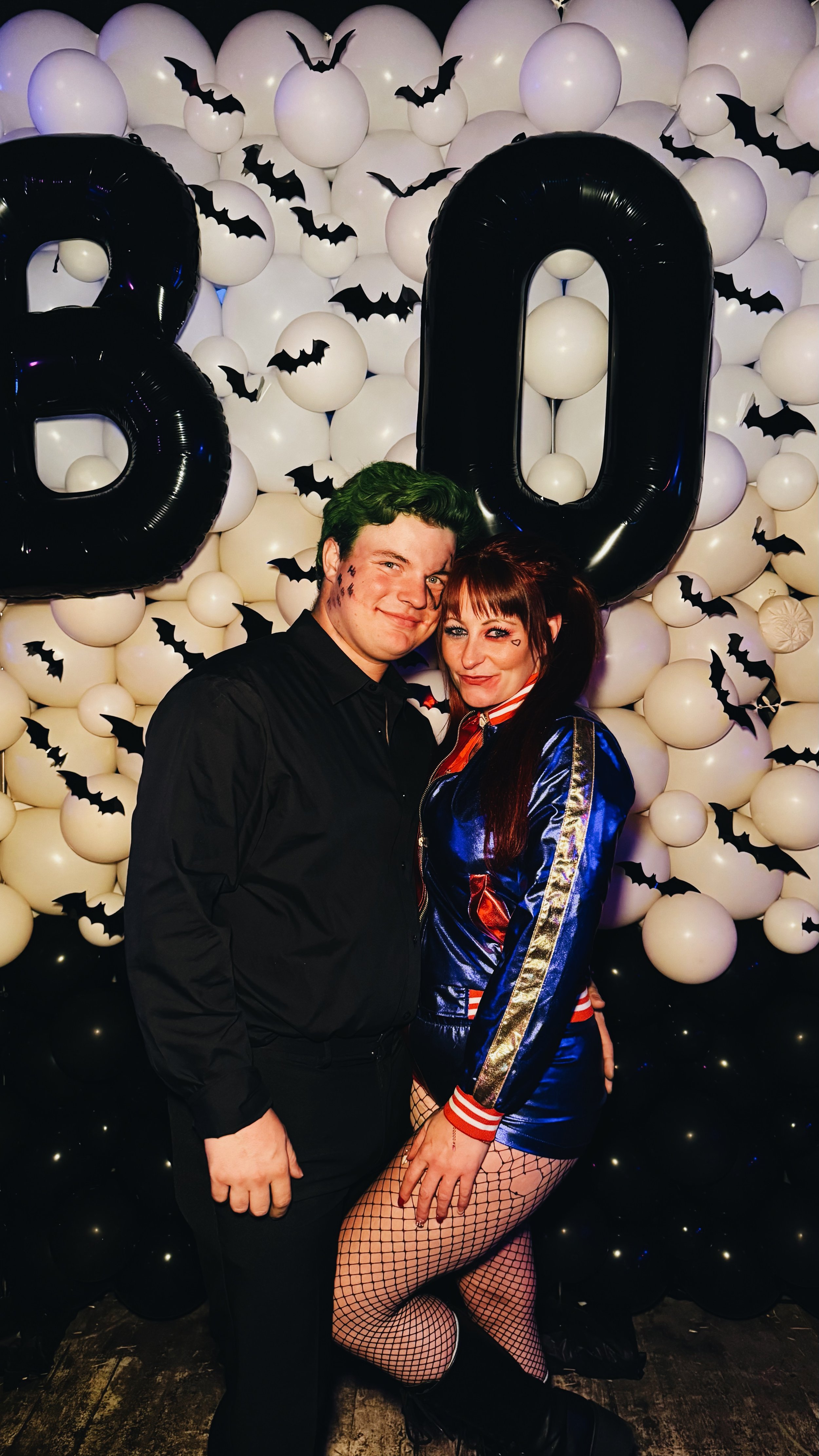 Two people dressed in costumes posing in front of a Halloween themed backdrop with white balloons, black bat cutouts, and large black balloons forming the number 20.