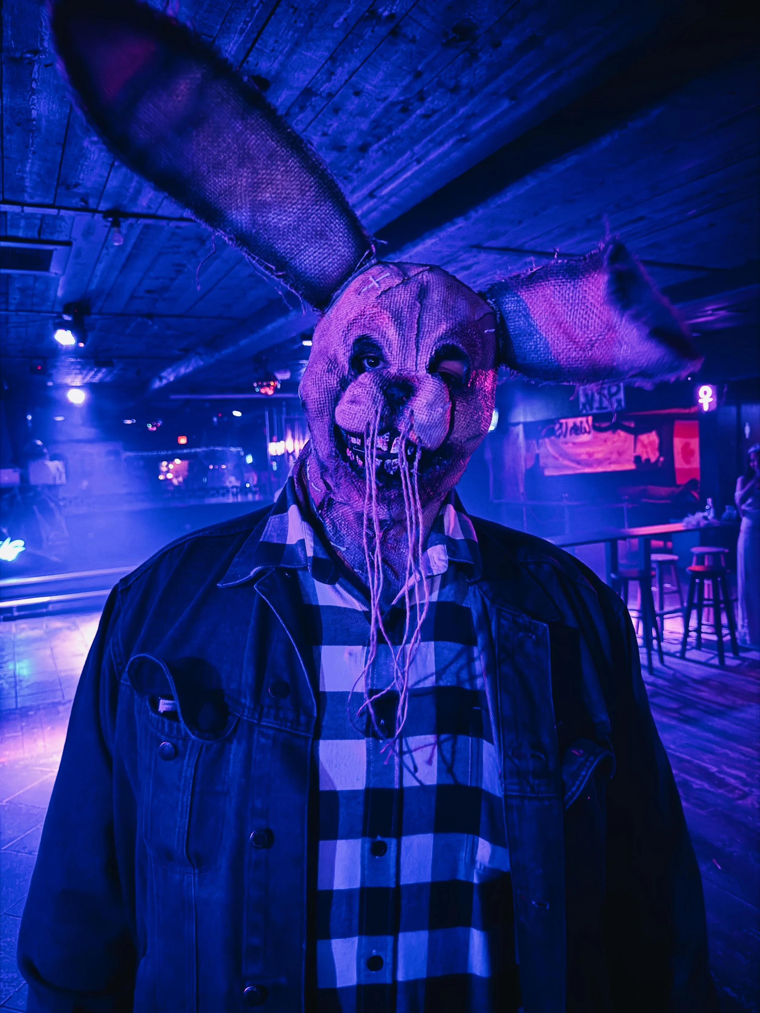 A person wearing a scary animal mask with large ears and a stitched face, dressed in a plaid shirt and jacket, in a dimly lit bar or club with neon lights.