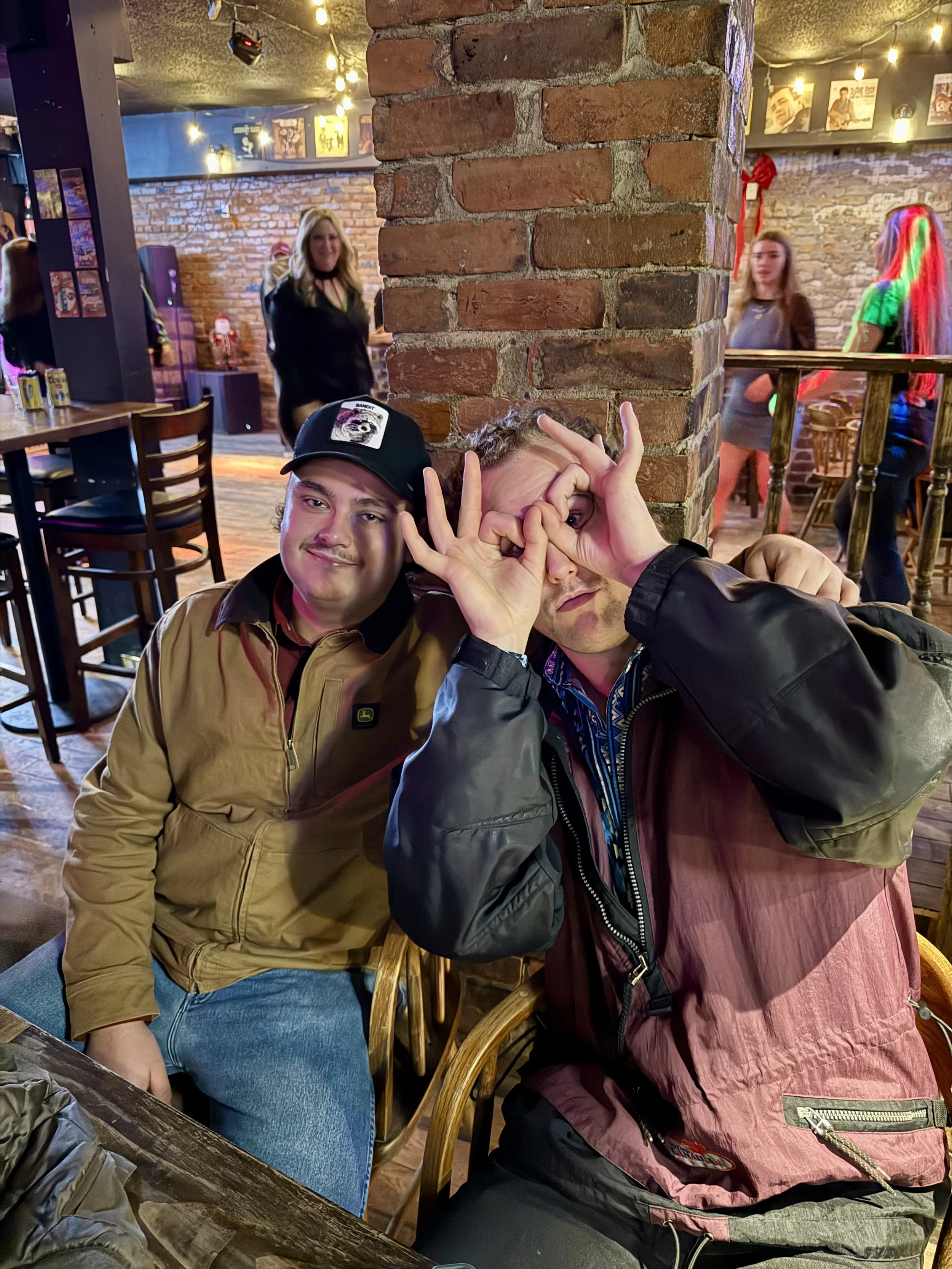 Two young men sitting at a table in a bar, with one making a playful binocular gesture with his hands around his eyes, and the other smiling at the camera. The bar has brick walls, string lights, and people in the background including women, one with