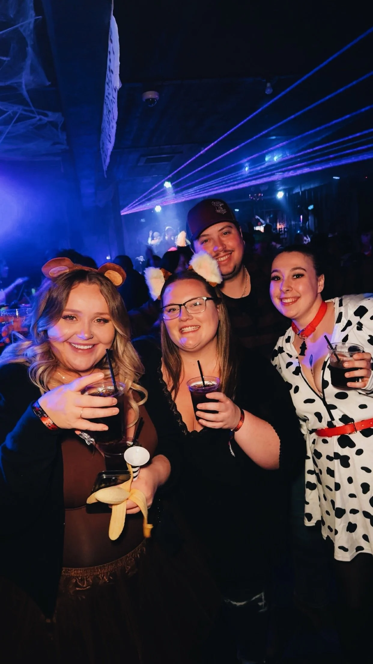 Group of four friends at a party, smiling and holding drinks, dressed in costume with cat ears, a cow print dress, and a baseball cap, surrounded by a lively nightclub scene with colorful laser lights.