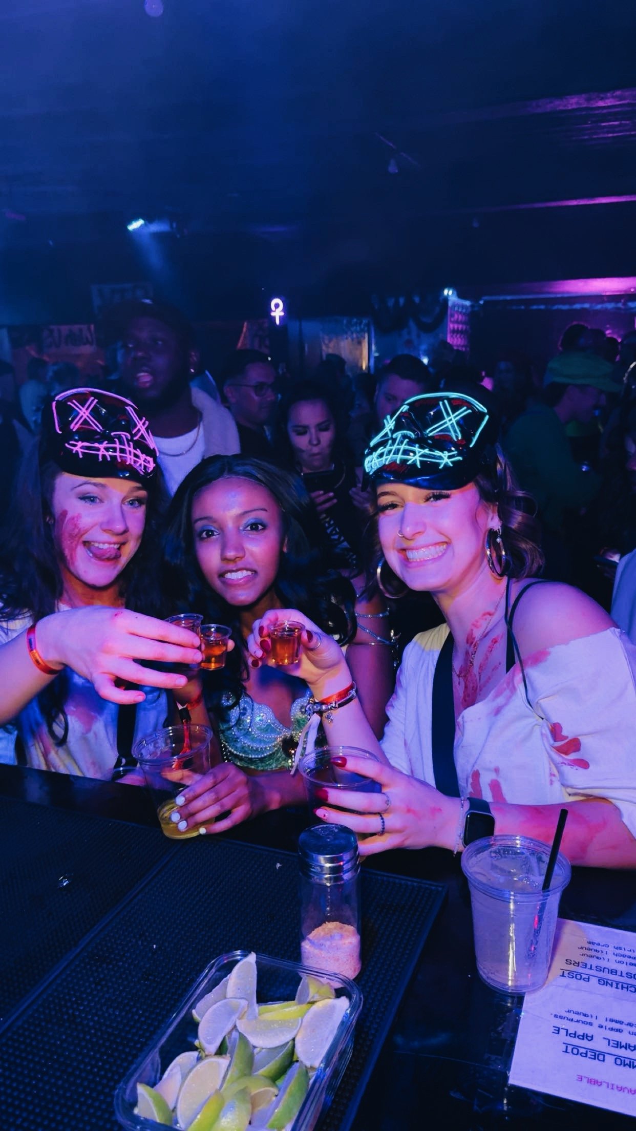 Three women celebrating at a party or nightclub, wearing LED hats, holding drinks, with a colorful, lively crowd in the background.