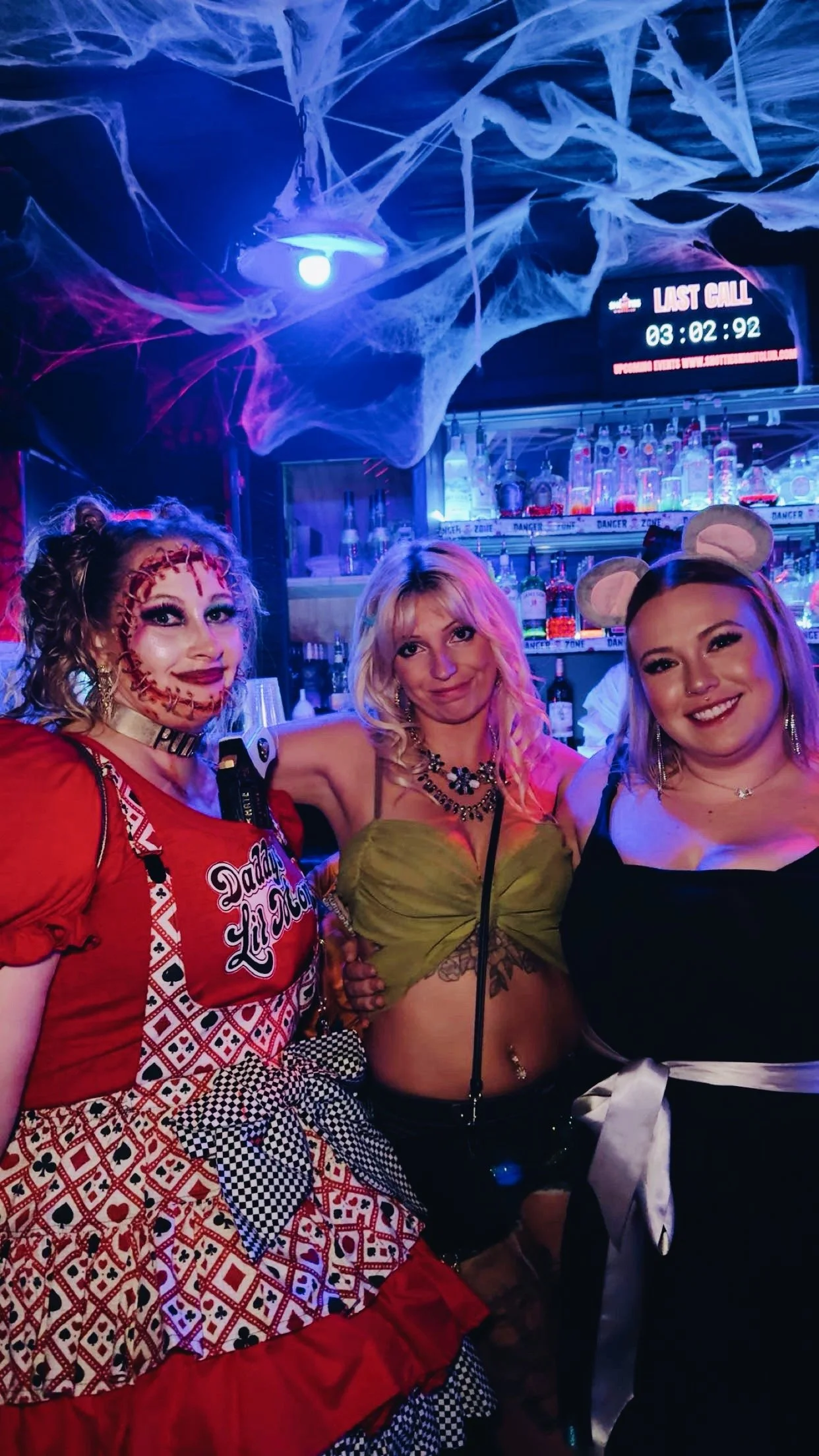 Three women in Halloween costumes at a bar decorated with cobwebs; one as a doll, another with mouse ears, and the third in a black dress with a satin belt.