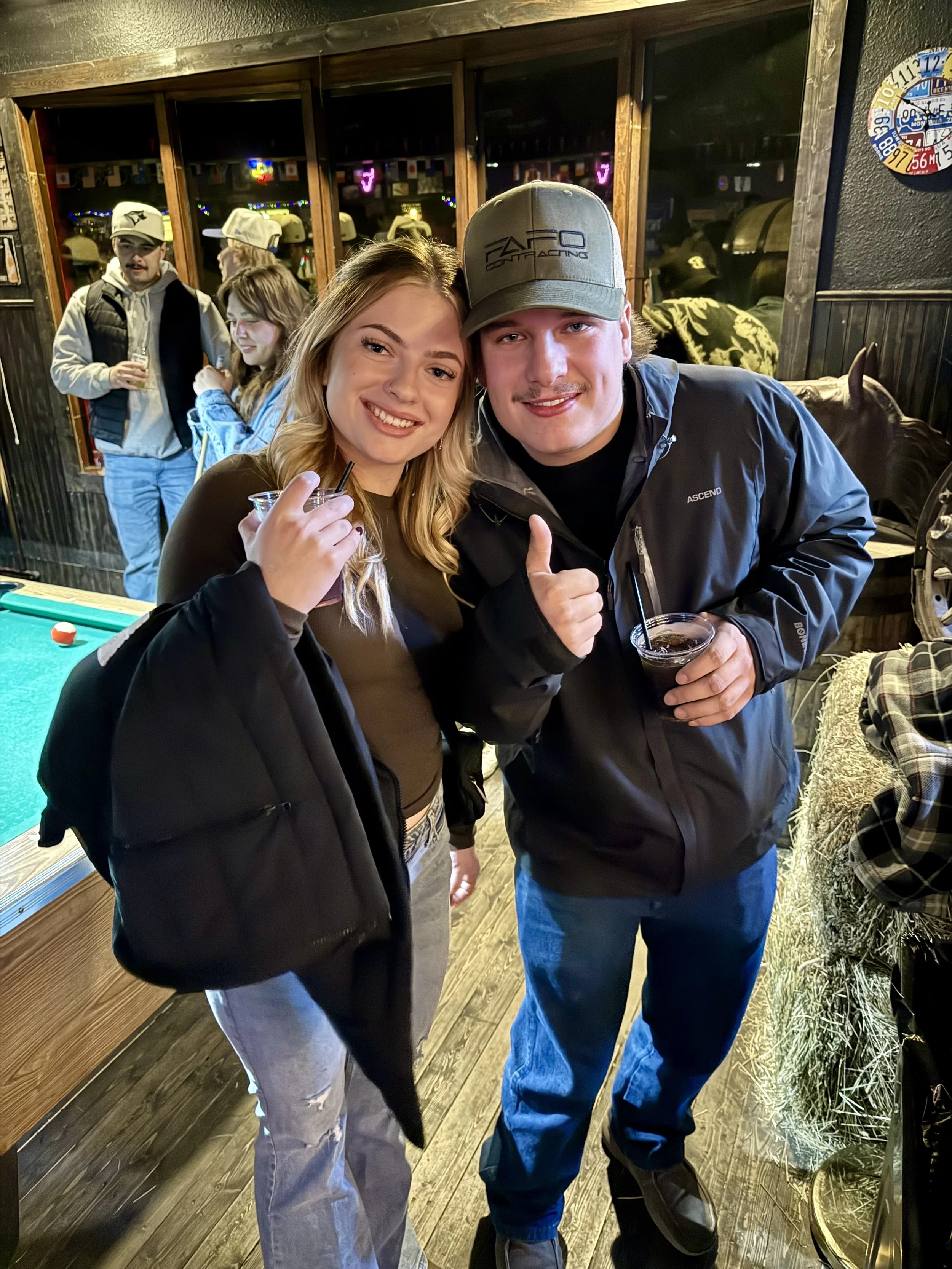 Two young adults, a woman and a man, smiling and holding drinks inside a bar or pub. They are standing close together, and the woman has a black jacket over her shoulder. The man is giving a thumbs-up and wearing a baseball cap. In the background, ot