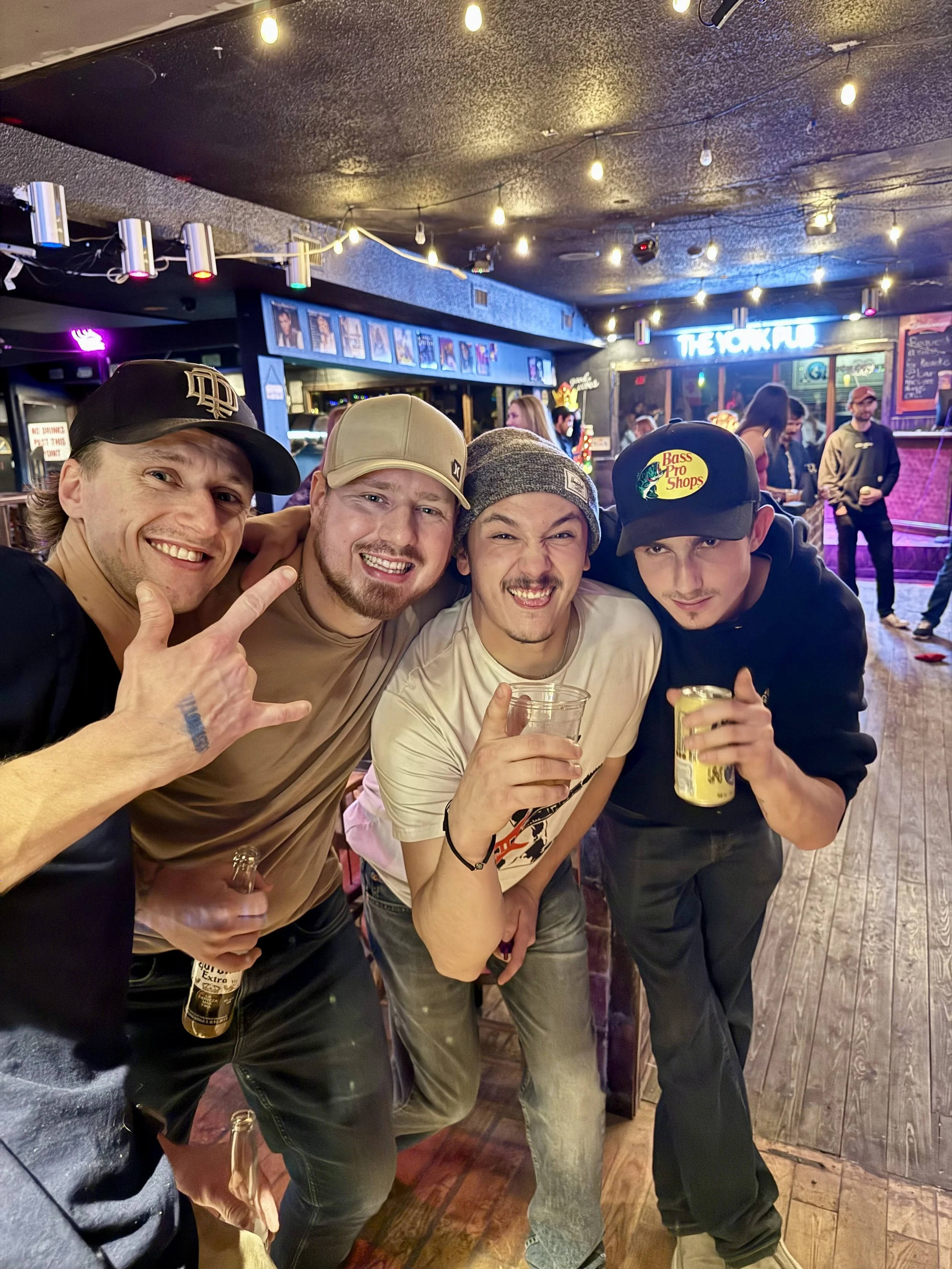 Four young men with casual clothing and hats enjoying drinks at a bar or concert venue, smiling and posing for the photo with colorful lights and other people in the background.