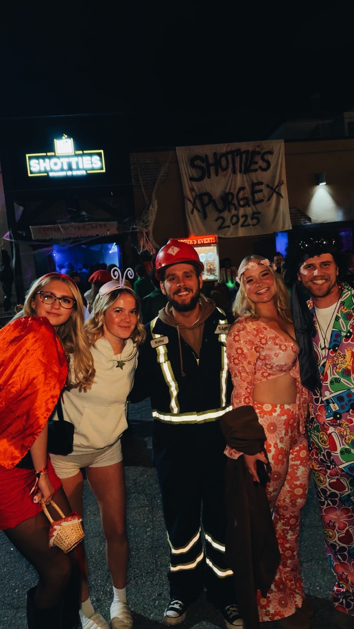Group of people in costumes at a Halloween event, with a sign that reads 'SHOTTIES XRRGEX 2025' in the background.