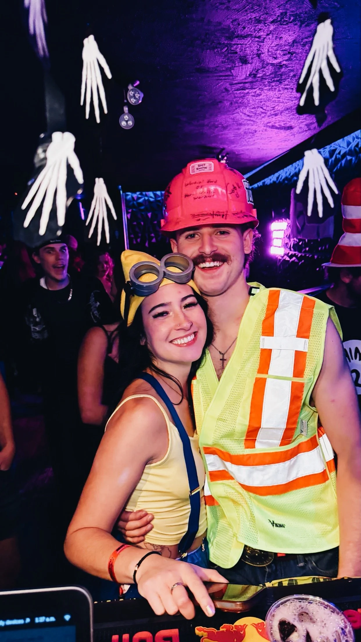 Two people dressed in costumes at a party, with skeleton decorations hanging from the ceiling. The man is wearing a construction hat and safety vest, while the woman is wearing goggles and cat ears.