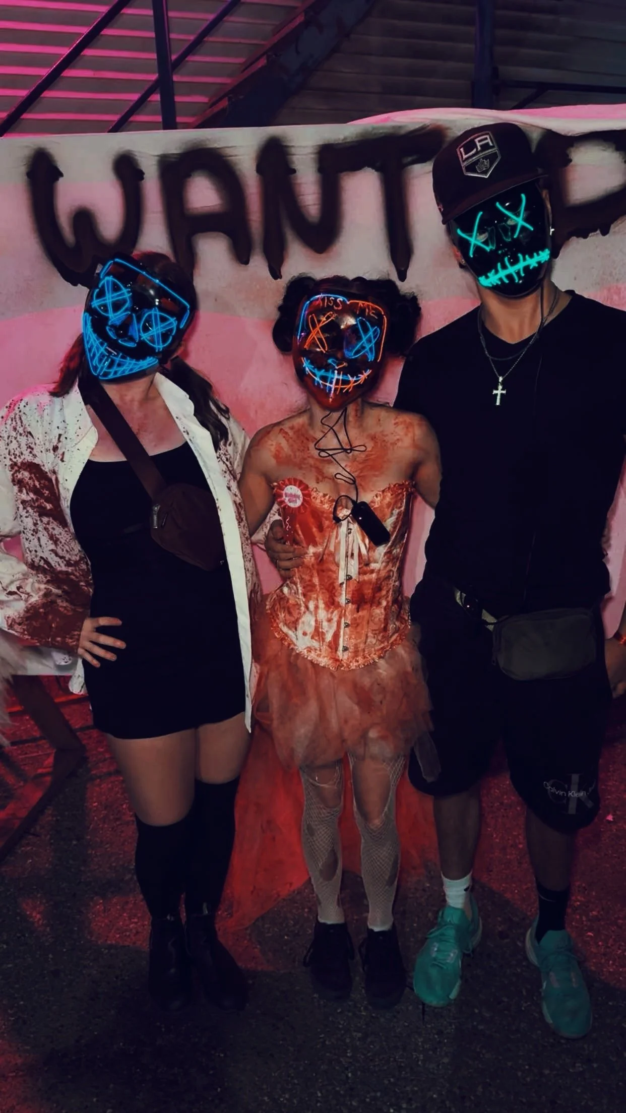 Three people wearing neon LED masks standing in front of a wall with black spray-painted letters that spell 'WANT'. The person in the middle is wearing an orange costume, while the other two are dressed in dark clothing.