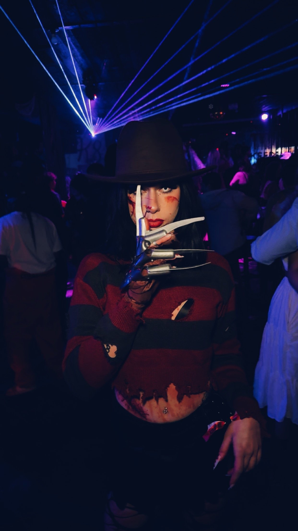 A woman dressed as Freddy Krueger from 'A Nightmare on Elm Street' at a costume party, with a fedora, striped sweater, and a Freddy glove, in a dimly lit nightclub with laser lights.