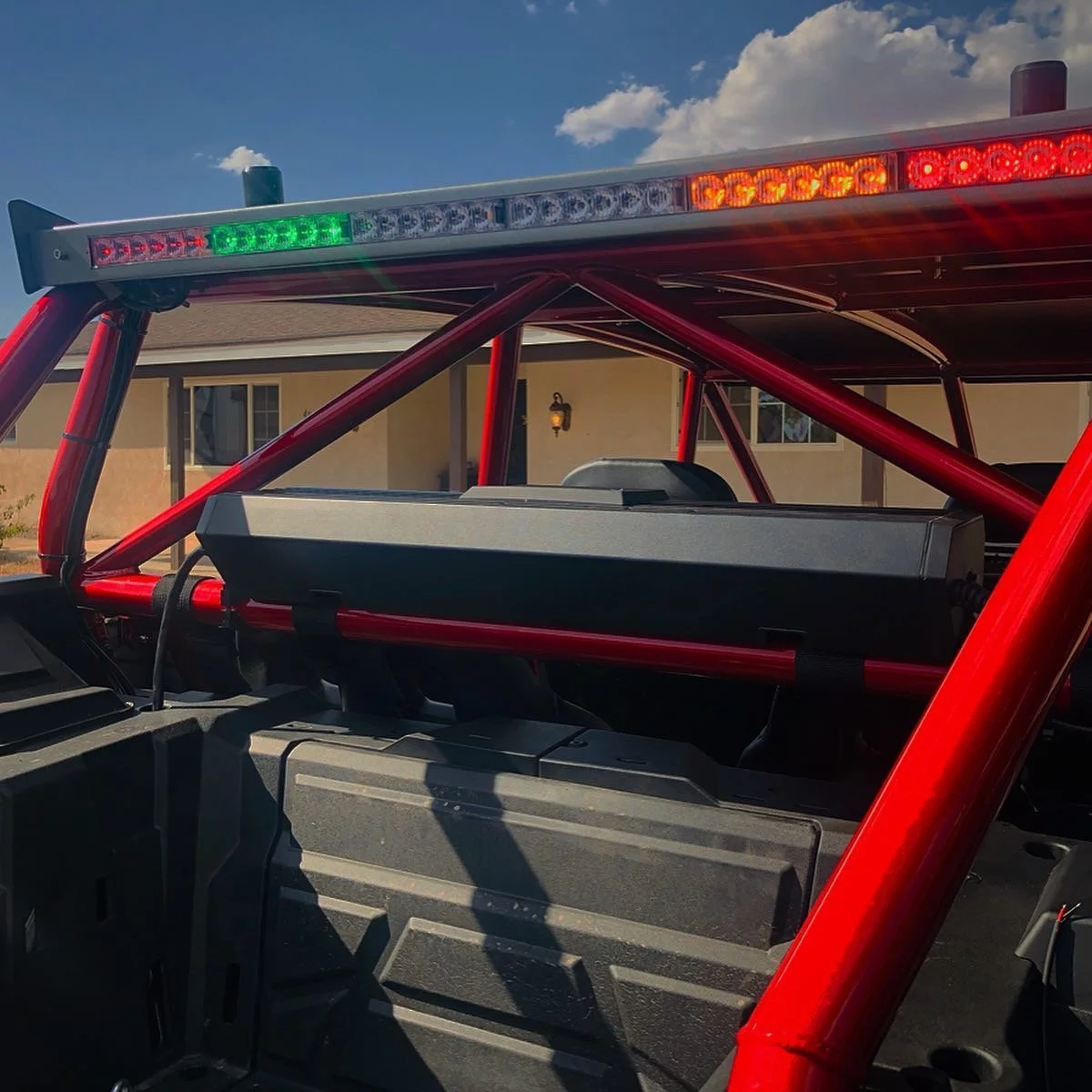 Close-up of a red off-road vehicle with a black roof rack and emergency lights on top, parked in front of a house under a blue sky with some clouds.