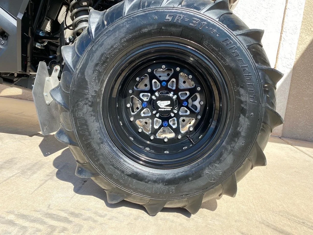 Close-up of off-road vehicle tire, labeled Bridgestone Dueler mud terrain with aggressive tread pattern, mounted on black rim with blue accents, part of a vehicle on a concrete surface.