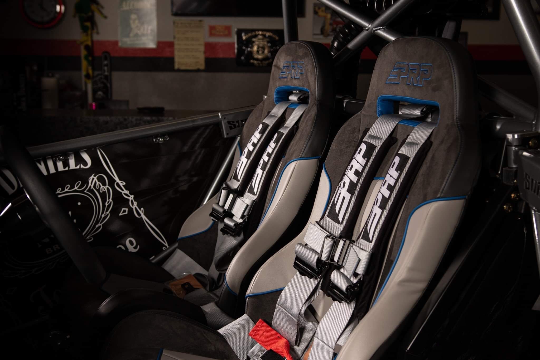 Two racing seats with safety harnesses inside a vehicle, with a background of posters and equipment on a garage wall.
