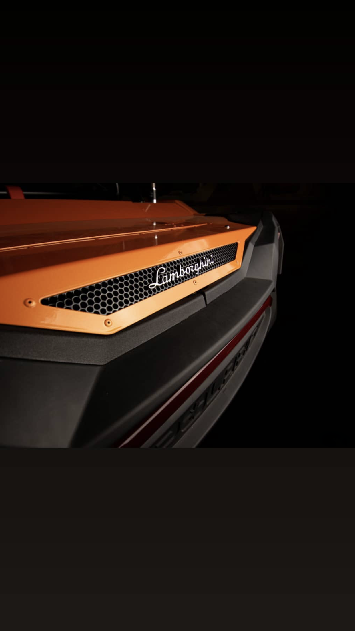 Close-up of a Lamborghini car with an orange hood and black grille, displaying the Lamborghini logo.