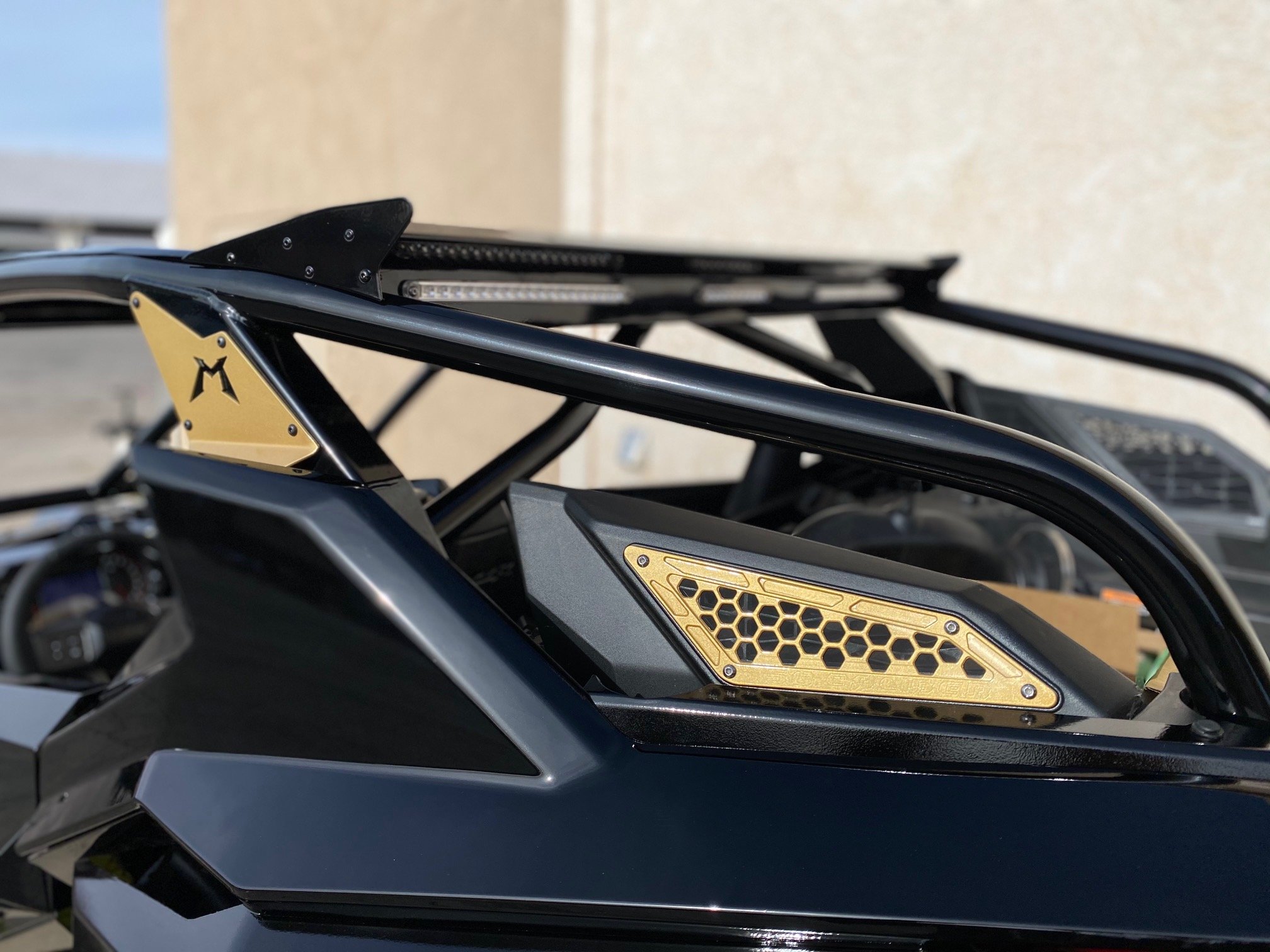 Close-up of a black and gold side panel on a snowmobile, showing a honeycomb vent design and the logo 'M' on the gold panel.