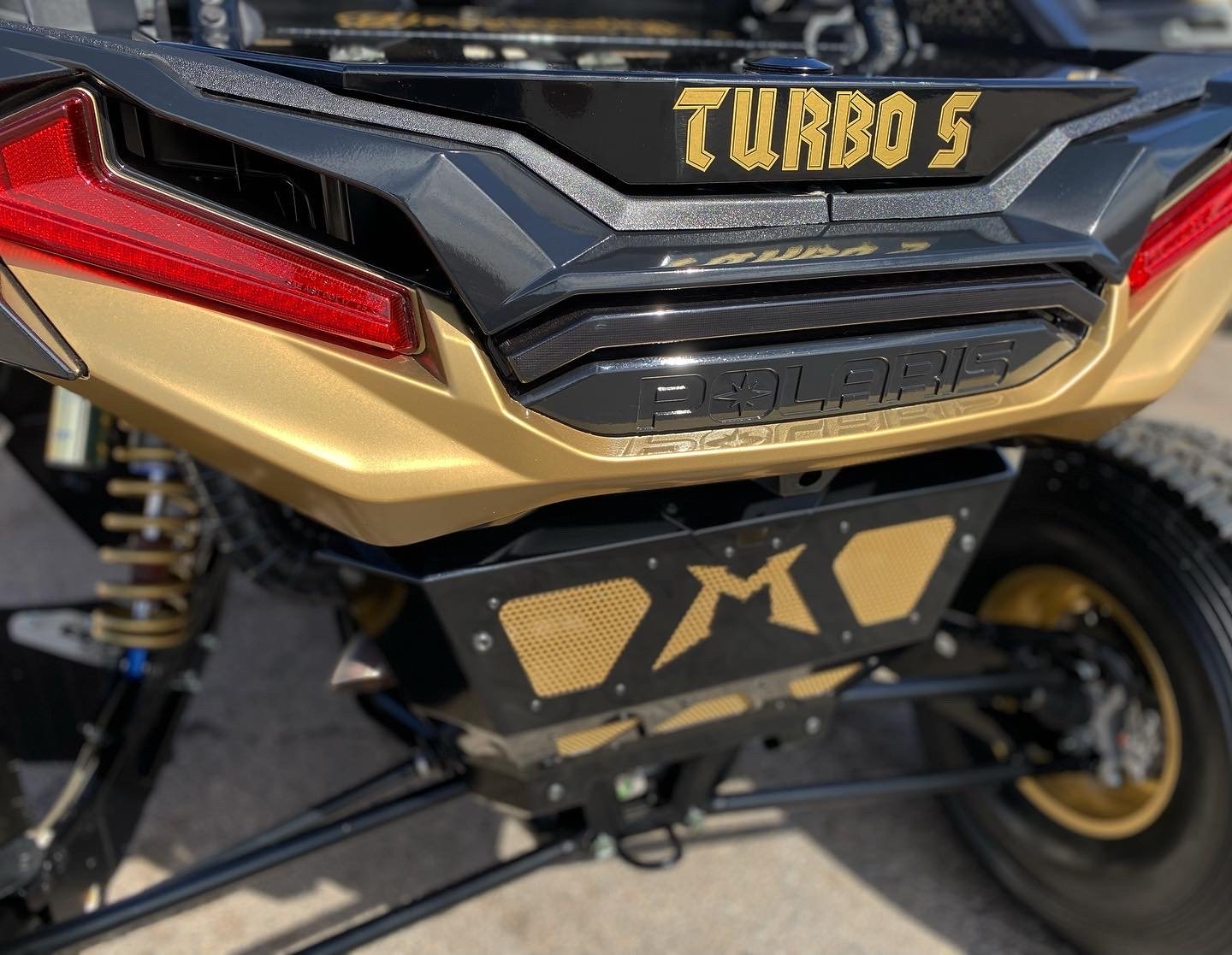 Close-up of a Polaris Turbo S snowmobile rear, featuring gold and black accents with decals that read 'TURBO S' and 'POLARIS', with a suspension and rear wheel visible.
