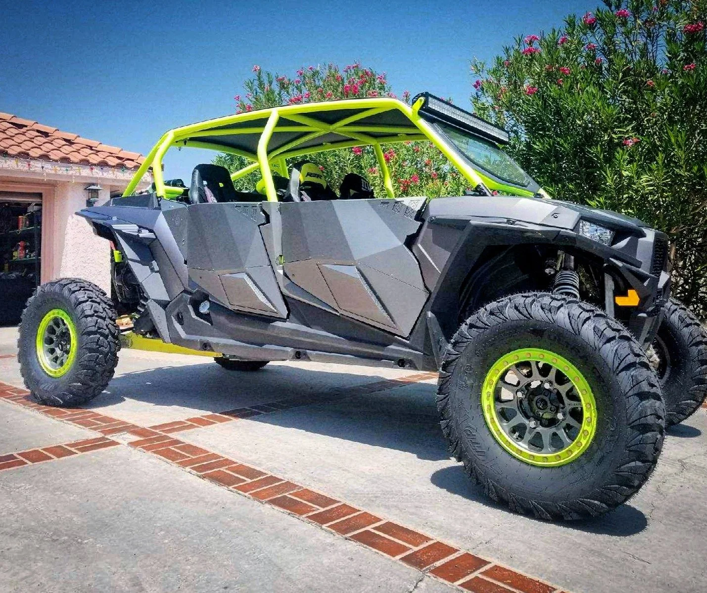 A custom off-road vehicle with a black body, neon yellow roll cage, large rugged tires, parked on a driveway in front of a house and green bushes.