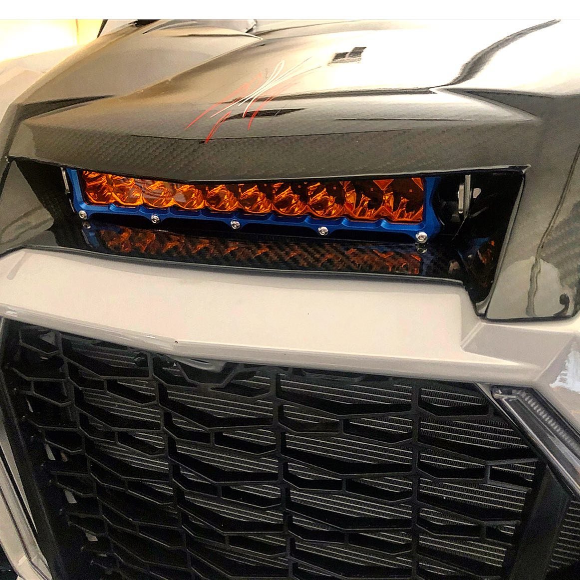 Close-up of the front of a vehicle with a black grille, carbon fiber hood, and custom LED headlights with an orange tint.
