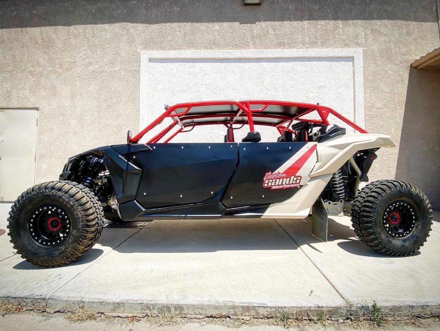 Side view of a custom off-road vehicle with large tires, black body panels, red roll cage, and a white front panel, parked on concrete in front of a beige wall.