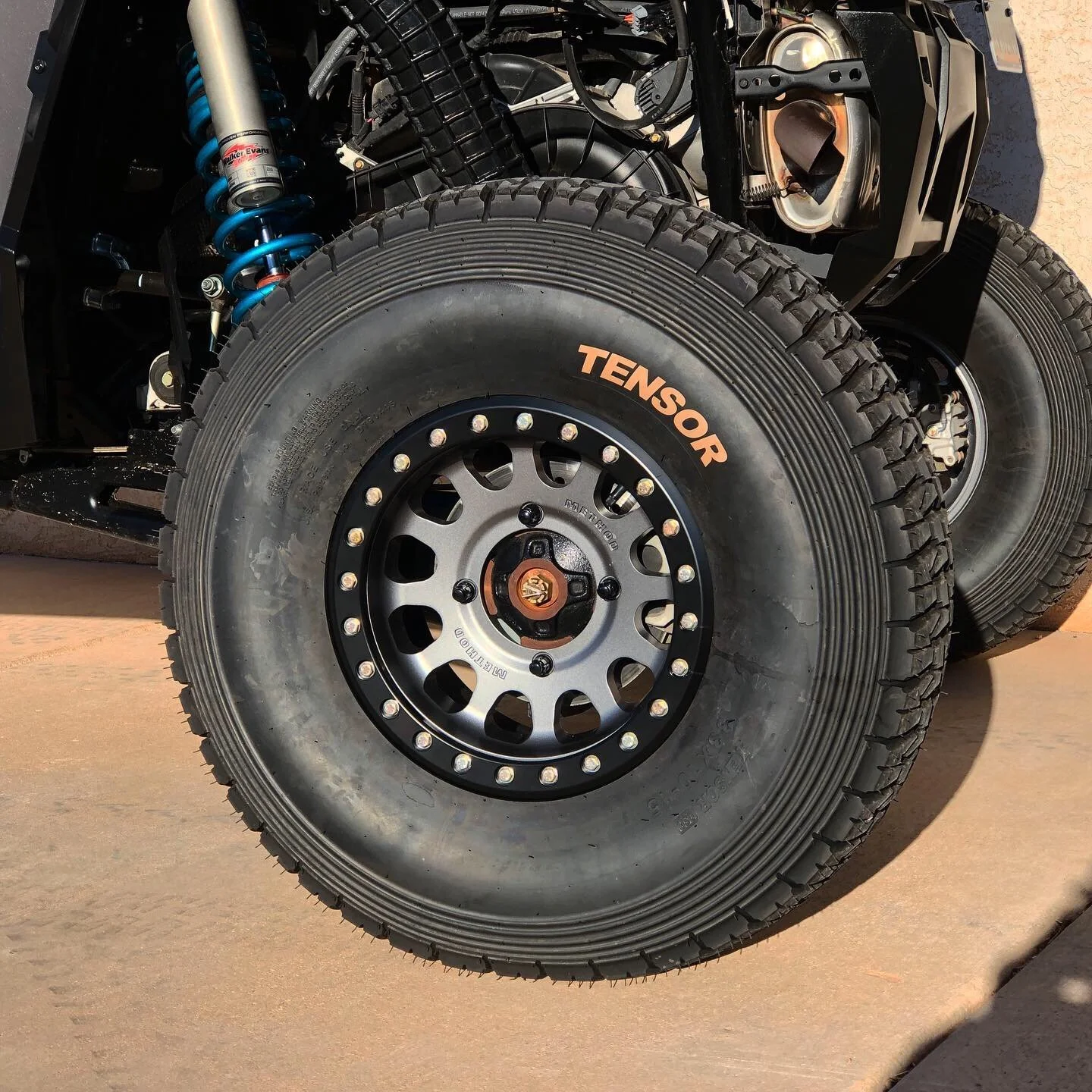 Close-up of off-road tire with the brand name TENSAR printed on the sidewall, mounted on a black wheel with a multi-hole pattern, attached to a vehicle with visible suspension components and partially exposed engine parts.