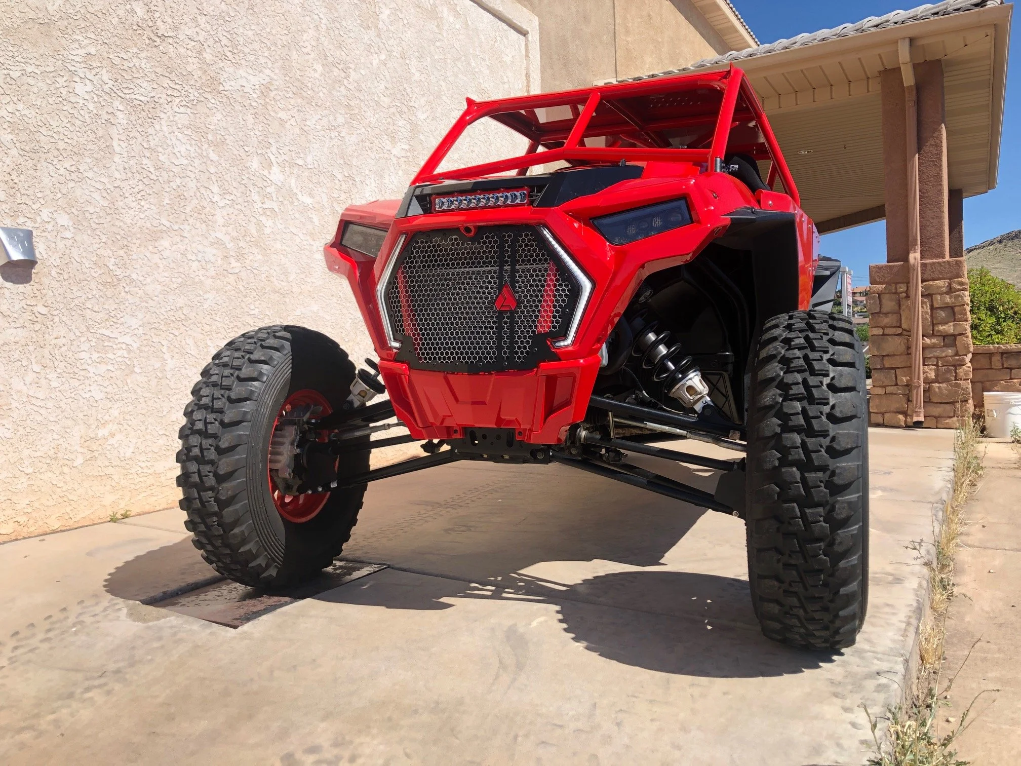 Red off-road vehicle with large tires, exposed suspension, parked on concrete driveway next to a house wall and porch.