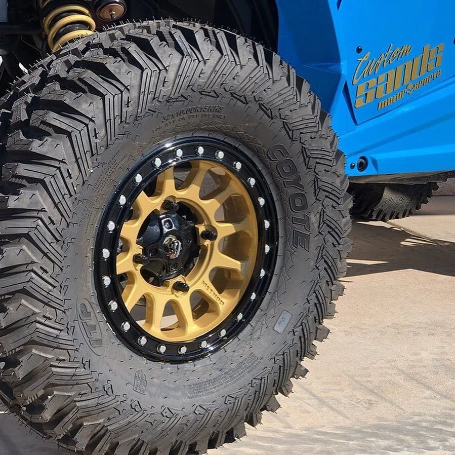 Close-up of a large off-road tire on a gold-colored wheel with black and silver accents. A blue vehicle body with gold lettering is partially visible in the background.