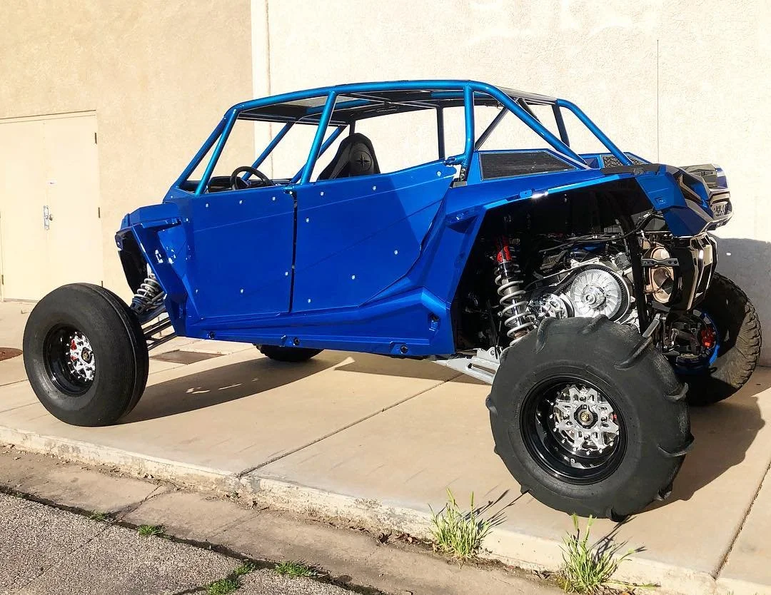 A partially assembled off-road vehicle chassis painted blue, with exposed wheels, suspension, and engine components, parked on a concrete driveway.