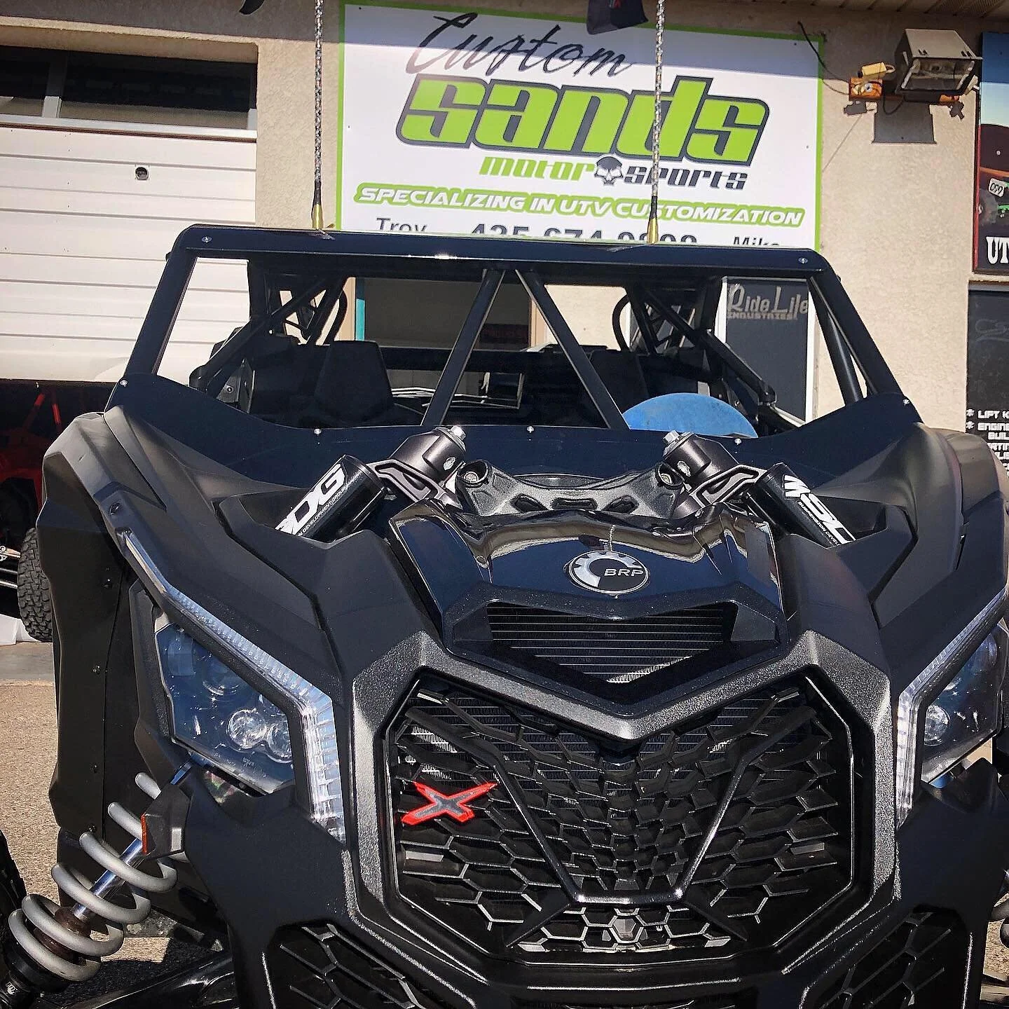Close-up of a black off-road vehicle with a prominent grille and Suspension, parked in front of a business sign that reads Custom Sans Motor Sports, specializing in custom off-road vehicles.