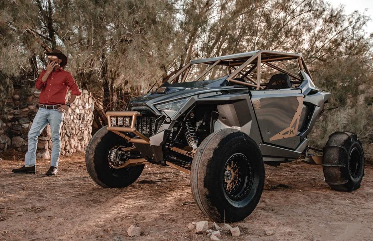 A rugged off-road vehicle with a roll cage and large tires parked on a dirt area, with a man in a cowboy hat and red shirt standing nearby, talking on a cell phone, in a dry, wooded environment.
