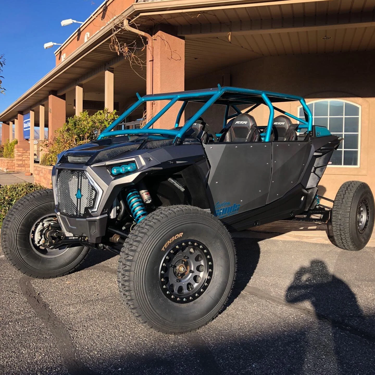 A custom off-road vehicle with large tires, blue roll cage, and black body panels parked outside a building.