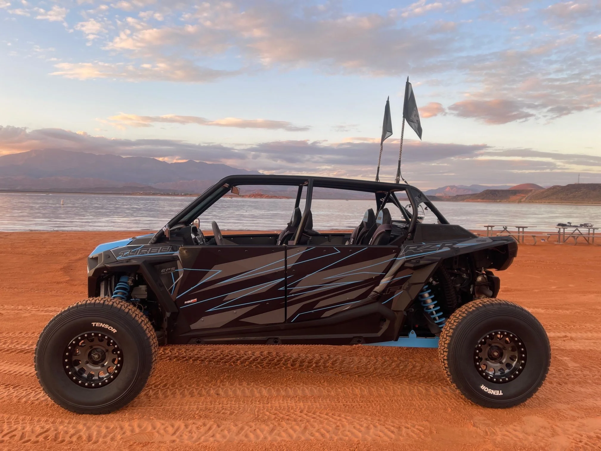 Off-road racing buggy parked on red dirt with mountains and lake in the background at sunset.