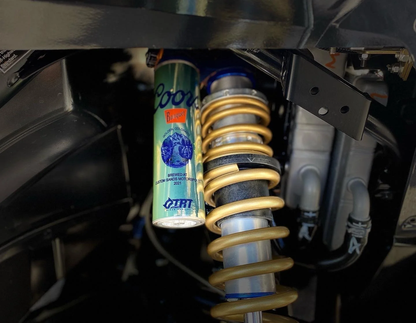 Close-up of a motorcycle or vehicle suspension system showing a coil shock absorber with a colorful can of Coors Banquet beer’s insulation or cover nearby.